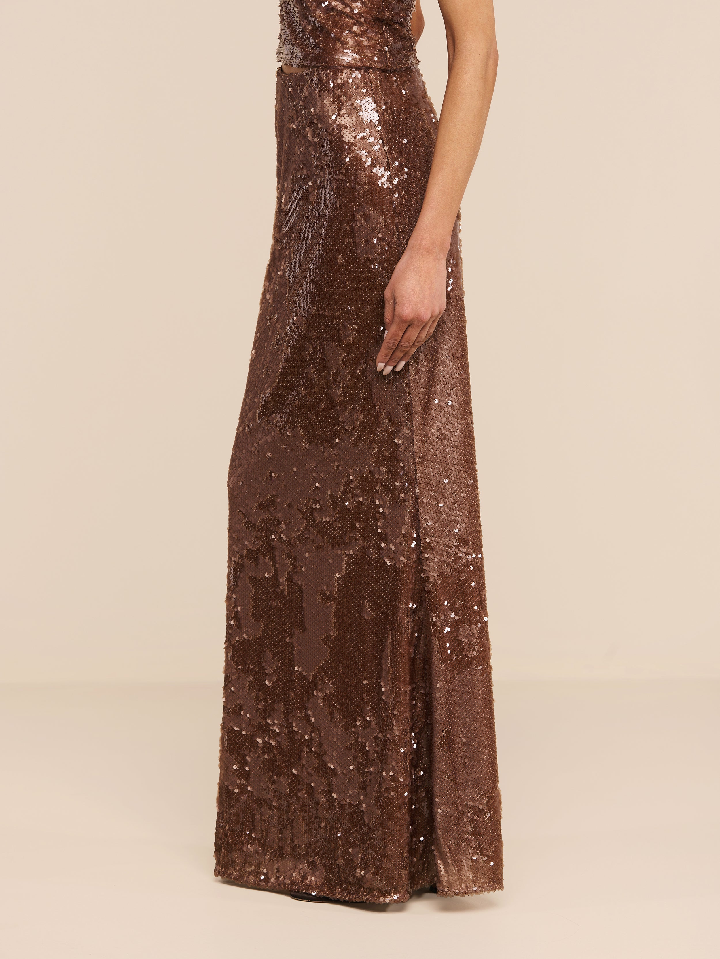 Una modella indossa la gonna lunga Zeta Sequin Maxi Skirt, un modello luccicante che arriva fino a terra della linea L’AGENCE, in piedi davanti a uno sfondo beige a tinta unita, con in vista solo la parte inferiore del corpo e un braccio.
