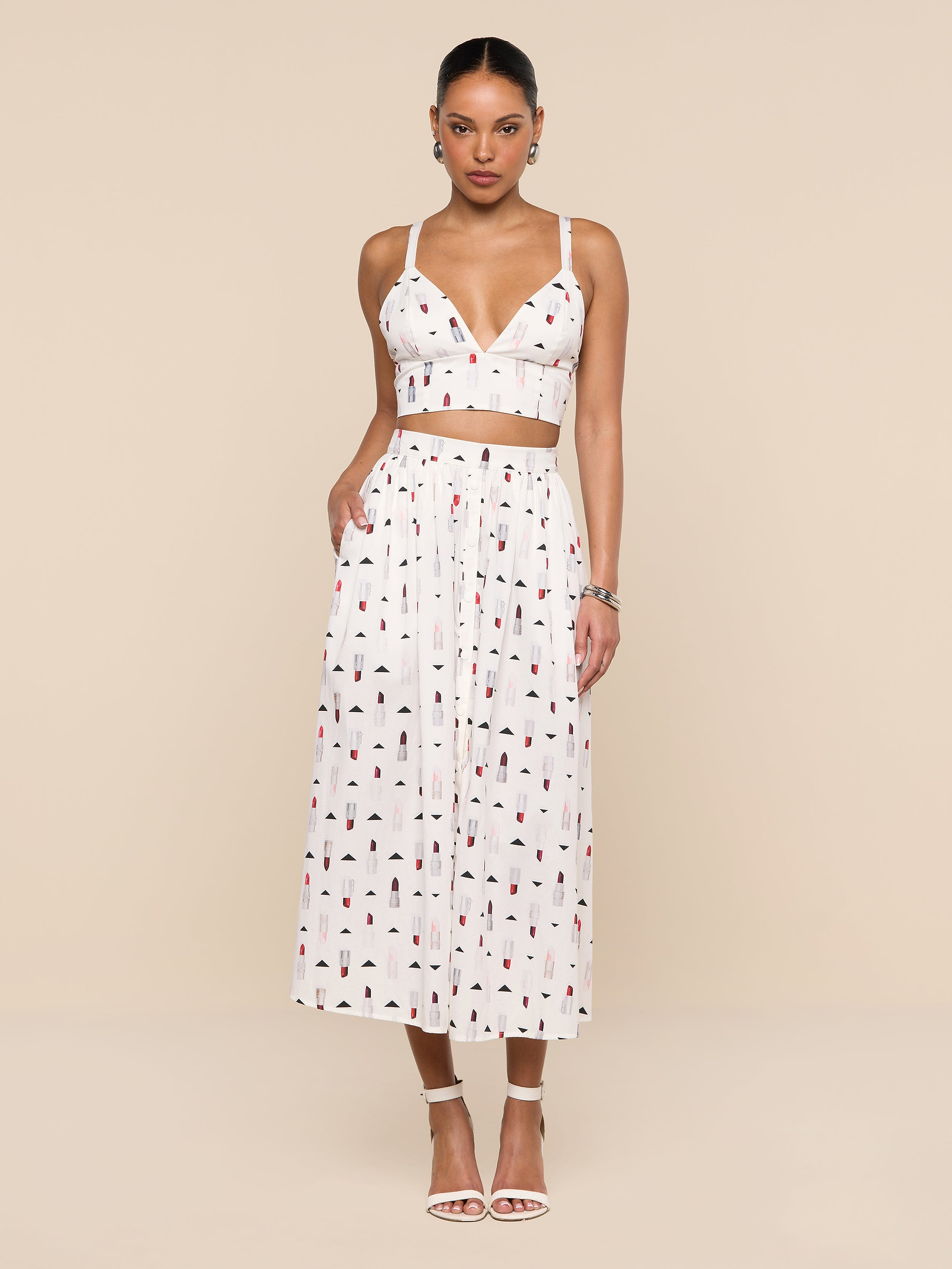 A woman stands against a white background wearing a white crop top and the Seina Poplin Midi Skirt—a high-waisted, ankle-length skirt featuring a small colorful print—paired with white high-heeled sandals.