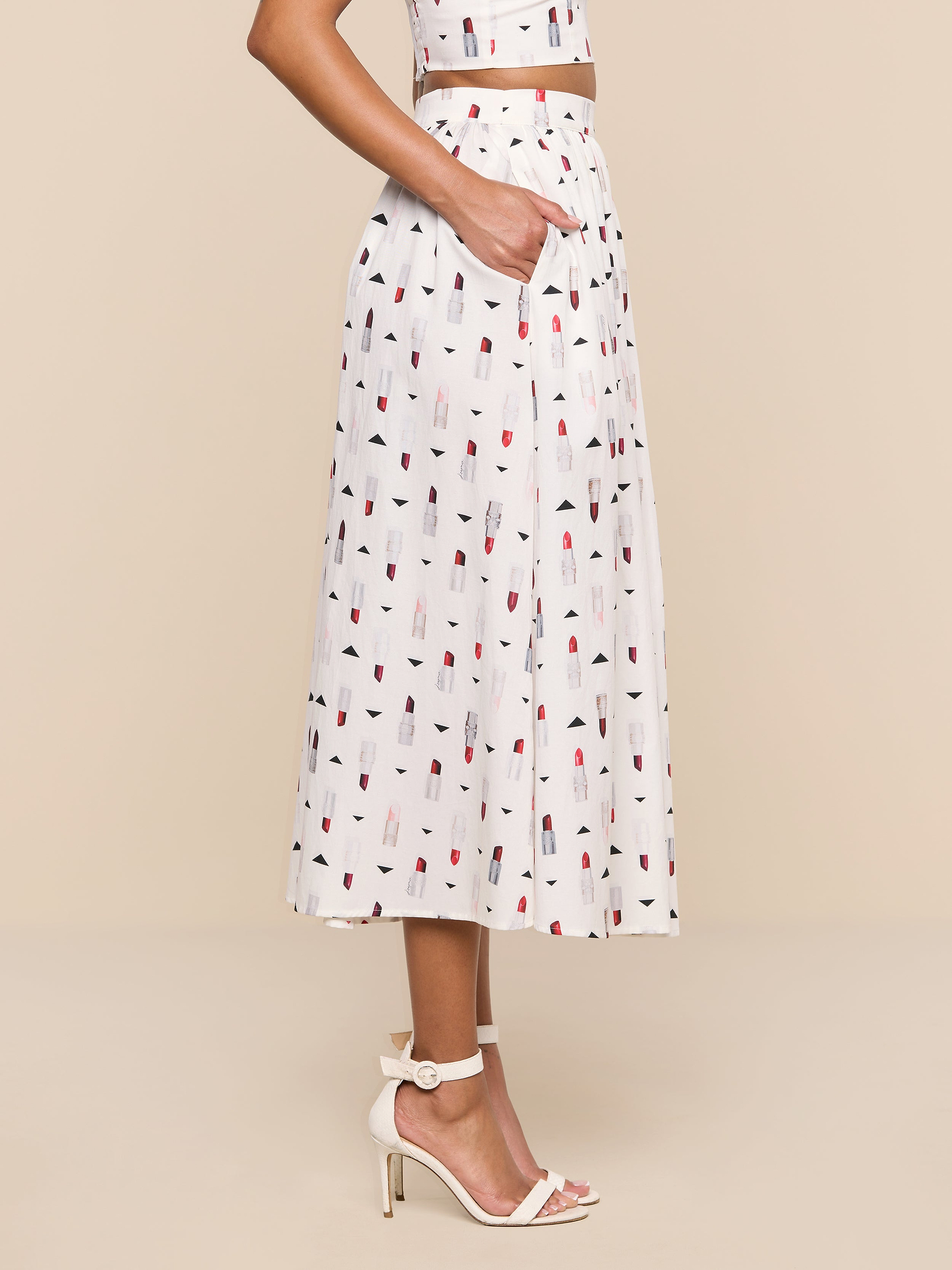 Sure Here’s a rewrite using the provided product name: A woman stands sideways against a white background, wearing the Seina Poplin Midi Skirt with a colorful lipstick print, paired with a matching crop top and blush high-heeled sandals, one hand tucked into her pocket.