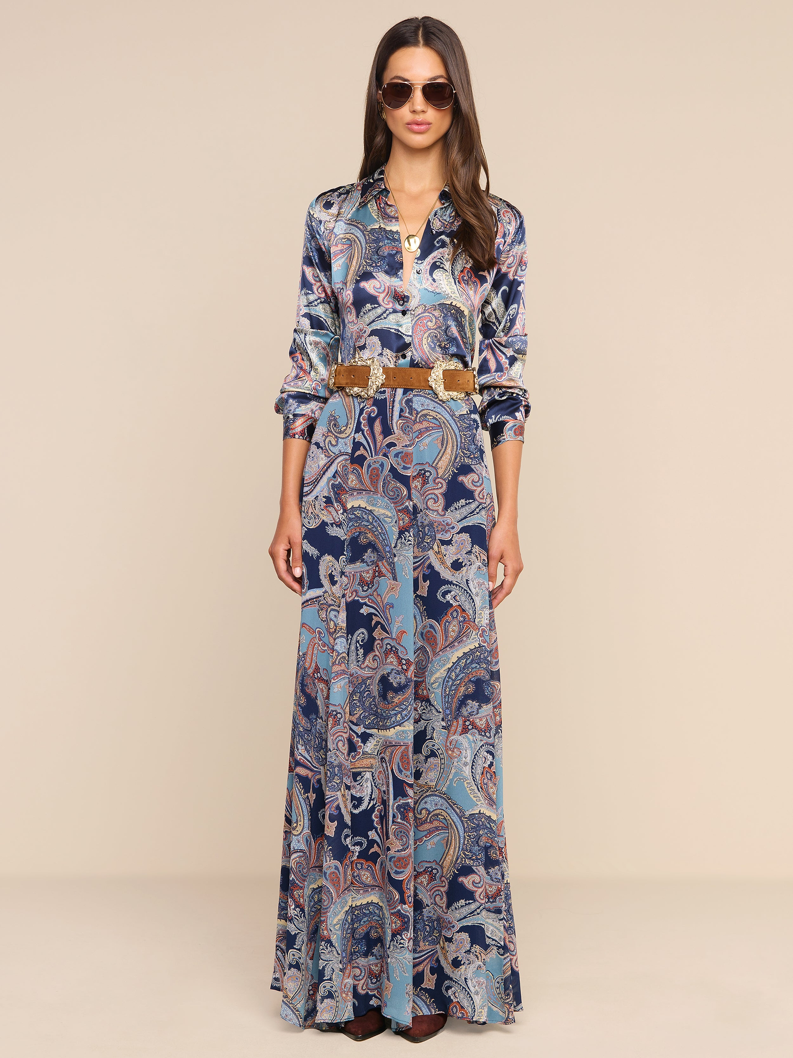 A woman wearing sunglasses and a long-sleeved top paired with the Patrica Maxi Skirt, featuring a floor-length paisley print and a brown belt, stands against a plain white background.