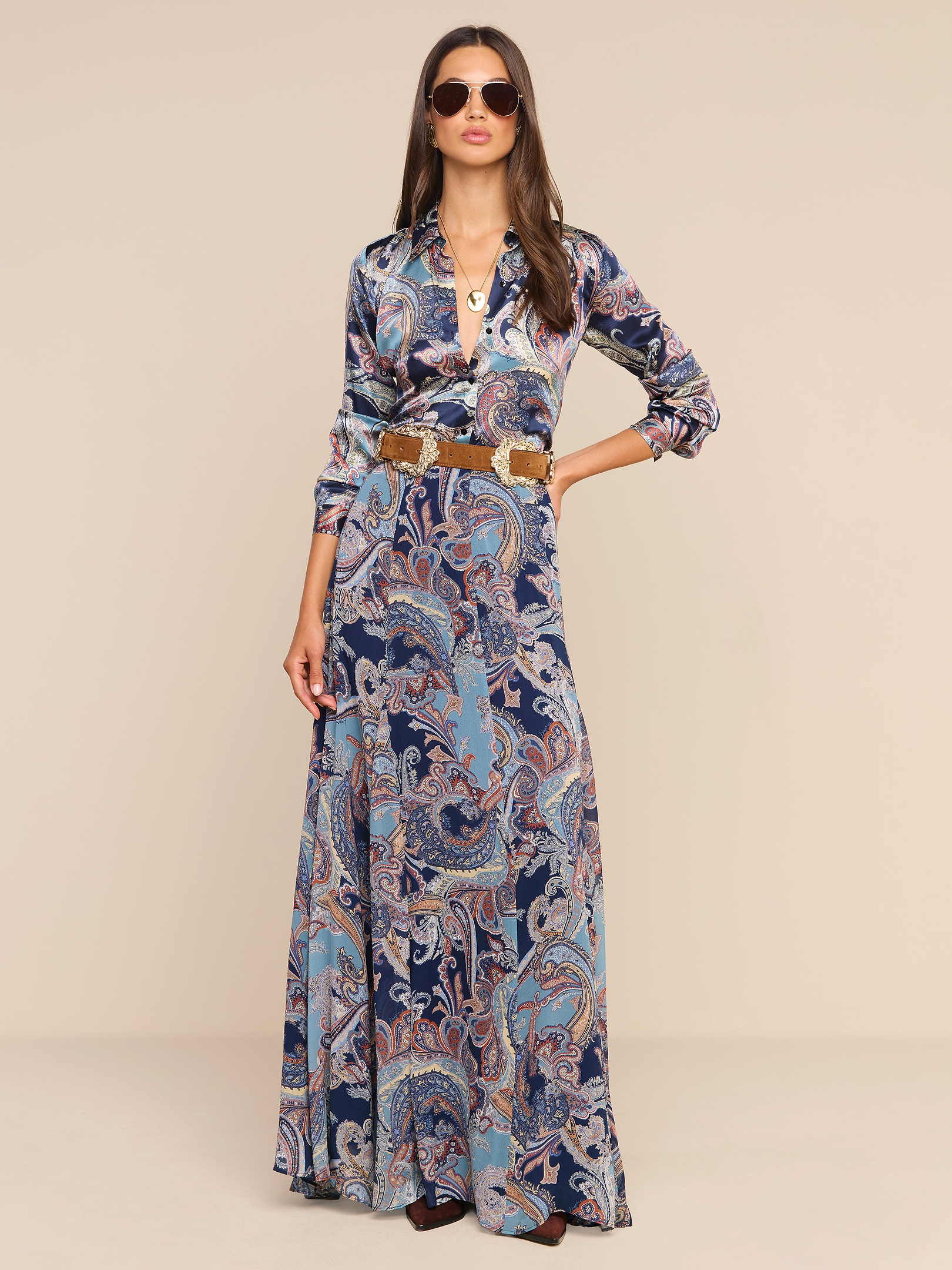 Una donna è in posa su uno sfondo beige a tinta unita, con indosso occhiali da sole e la gonna lunga Patrica Maxi Skirt: un modello che sfiora il pavimento con stampa paisley nei toni del blu e dell’arancione, impreziosito da delicati tocchi di blu notte che ne esaltano la profondità. La abbina a un top a maniche lunghe, una cintura larga e collane sovrapposte per completare il suo look di grande effetto.