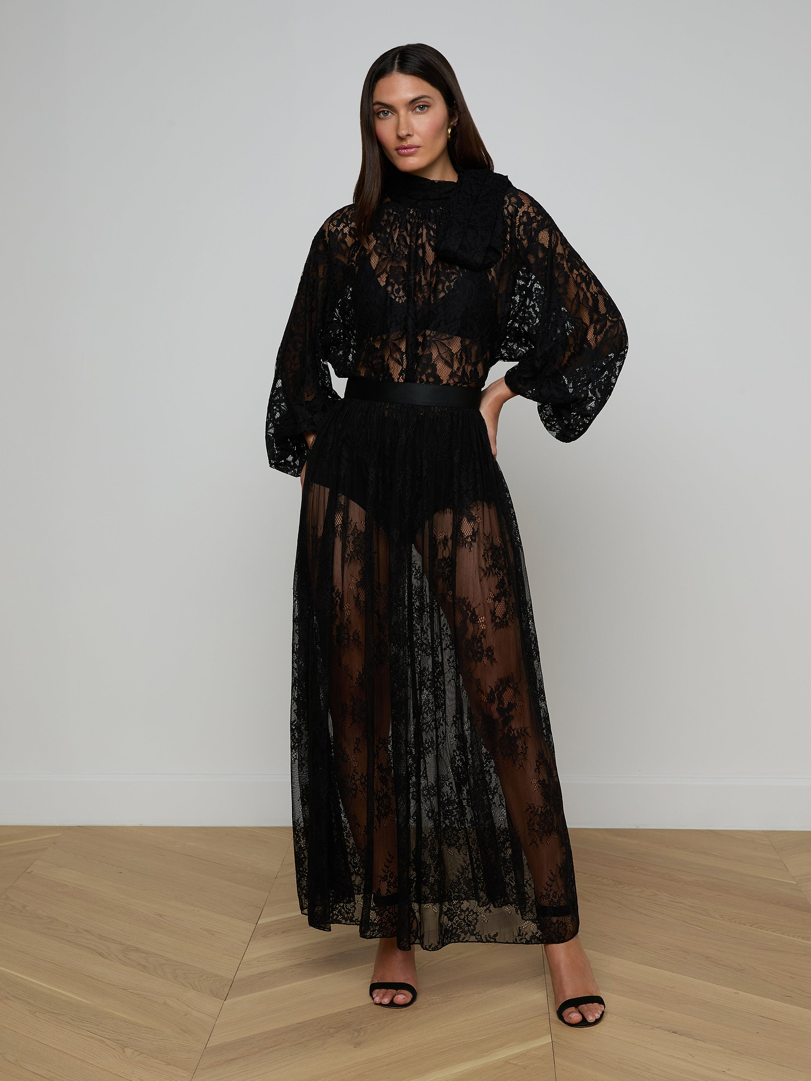 A woman stands against a plain white background wearing the Alona Lace Maxi Skirt in sheer black Chantilly floral lace, paired with a high-neck top featuring puffy sleeves and black high-heeled sandals.