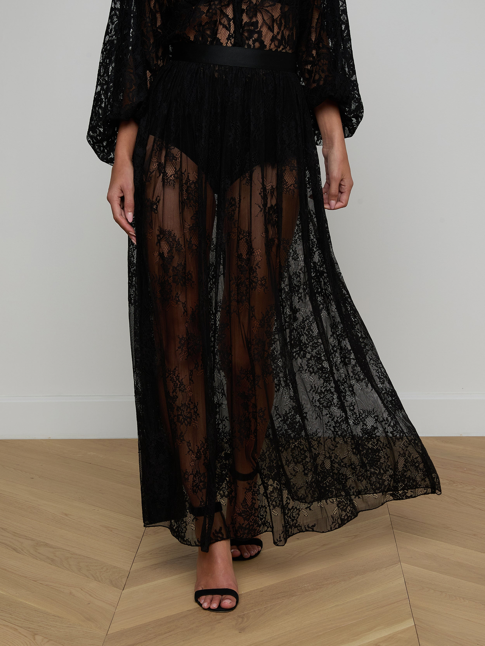Wearing the Alona Lace Maxi Skirt—a long, sheer black skirt featuring Chantilly floral lace—paired with black undergarments and black high-heeled sandals, this person stands against a plain light background.