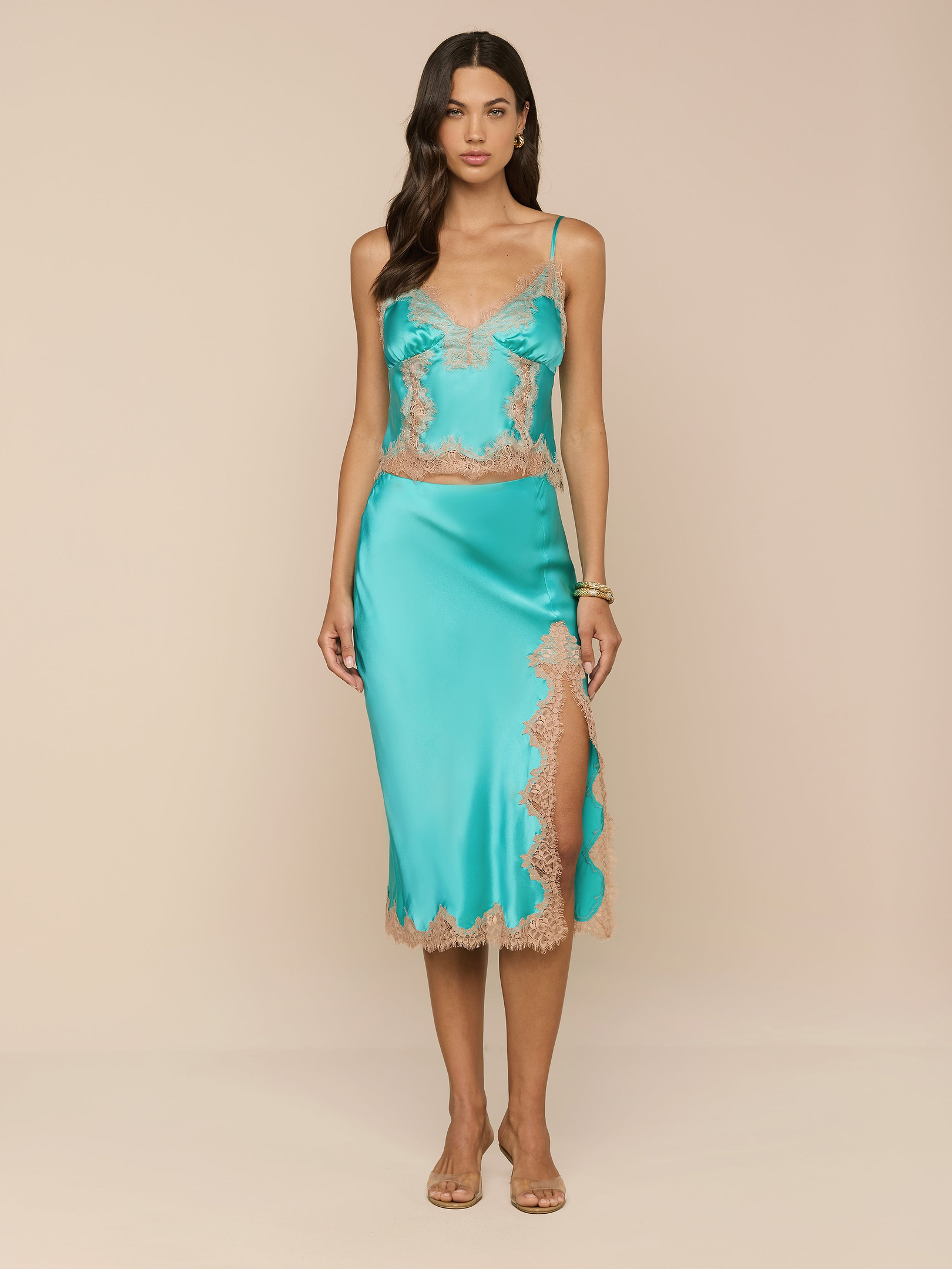 A woman stands facing forward wearing the Tico Silk Lace Skirt in teal with eyelash lace trim and a side slit, paired with nude heels, against a plain white background.