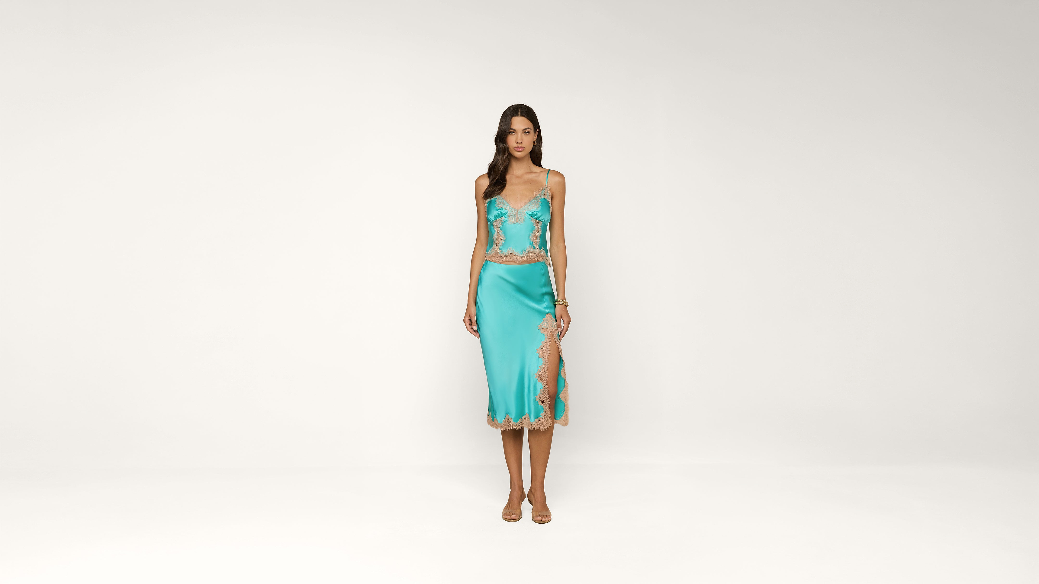 A woman stands against a plain white background wearing a turquoise lace-trimmed camisole and matching midi skirt with a slit, and beige heeled sandals.