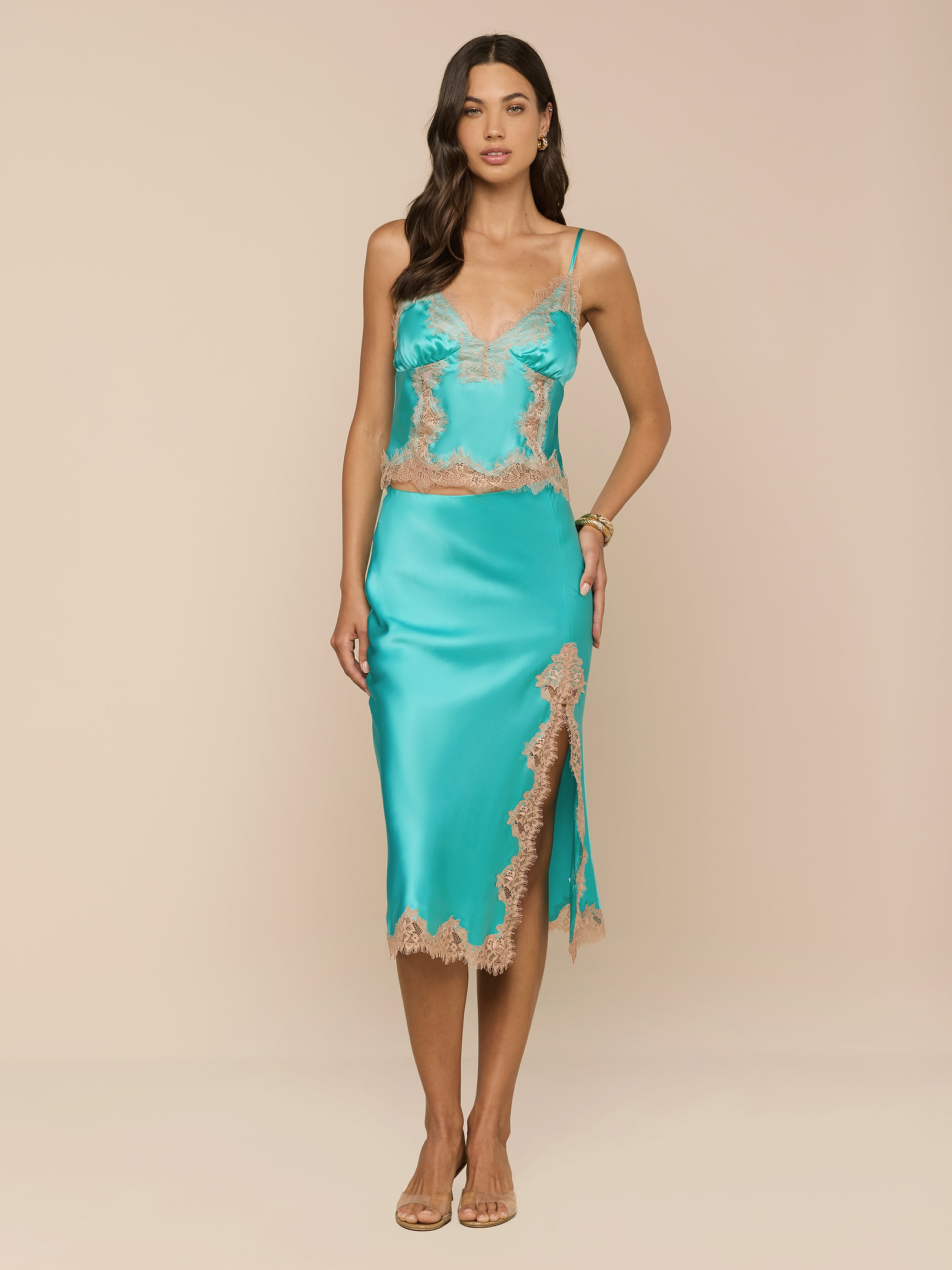A woman stands against a plain white background, wearing the Tico Silk Lace Skirt in turquoise satin with eyelash lace trim and a side slit, paired with sandals.