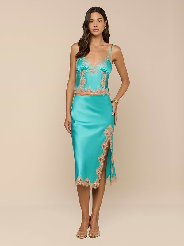 A woman stands against a plain white background, wearing the Tico Silk Lace Skirt in turquoise satin with eyelash lace trim and a side slit, paired with sandals.