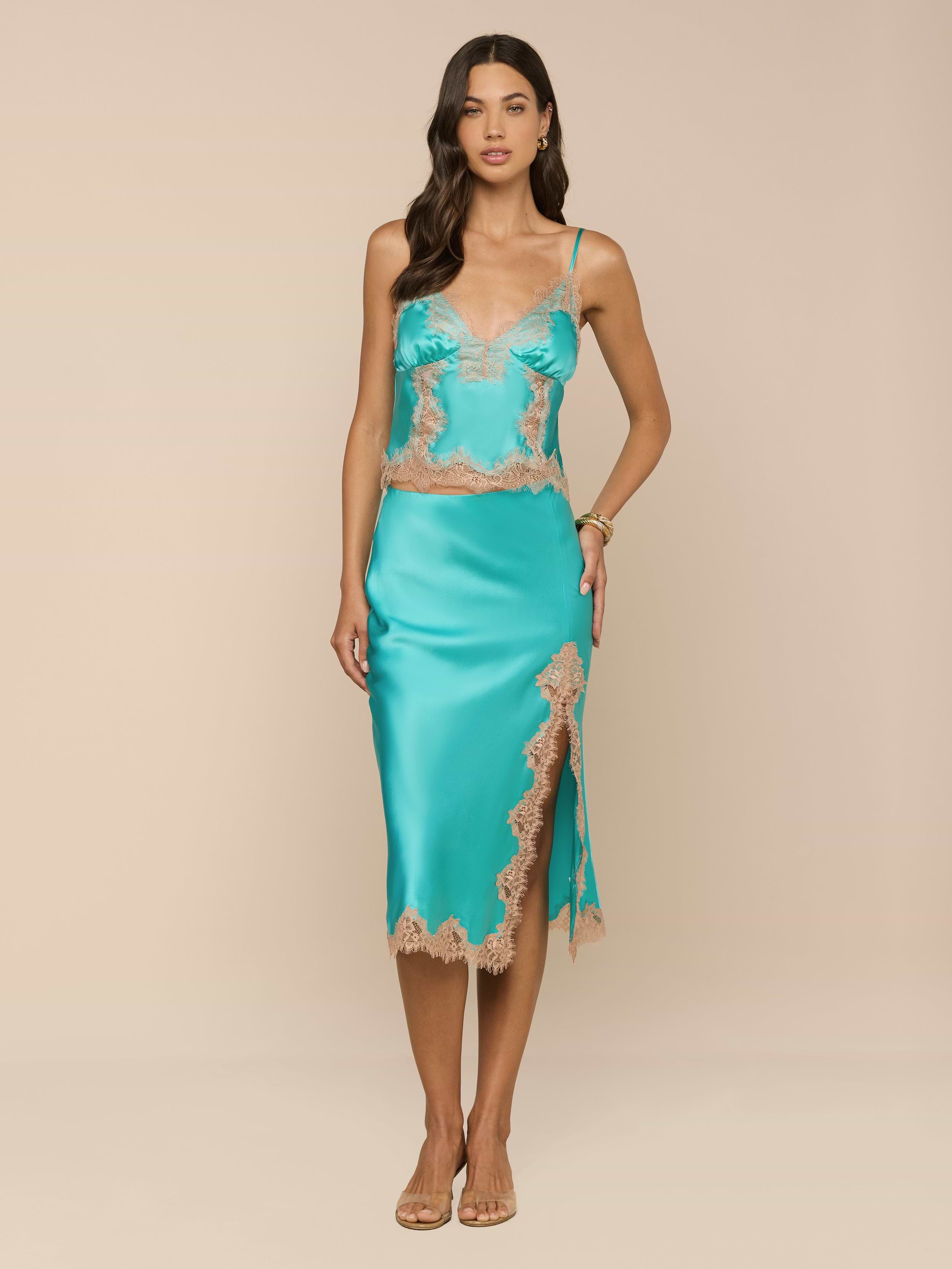 A woman stands against a plain white background, wearing the Tico Silk Lace Skirt in turquoise satin with eyelash lace trim and a side slit, paired with sandals.