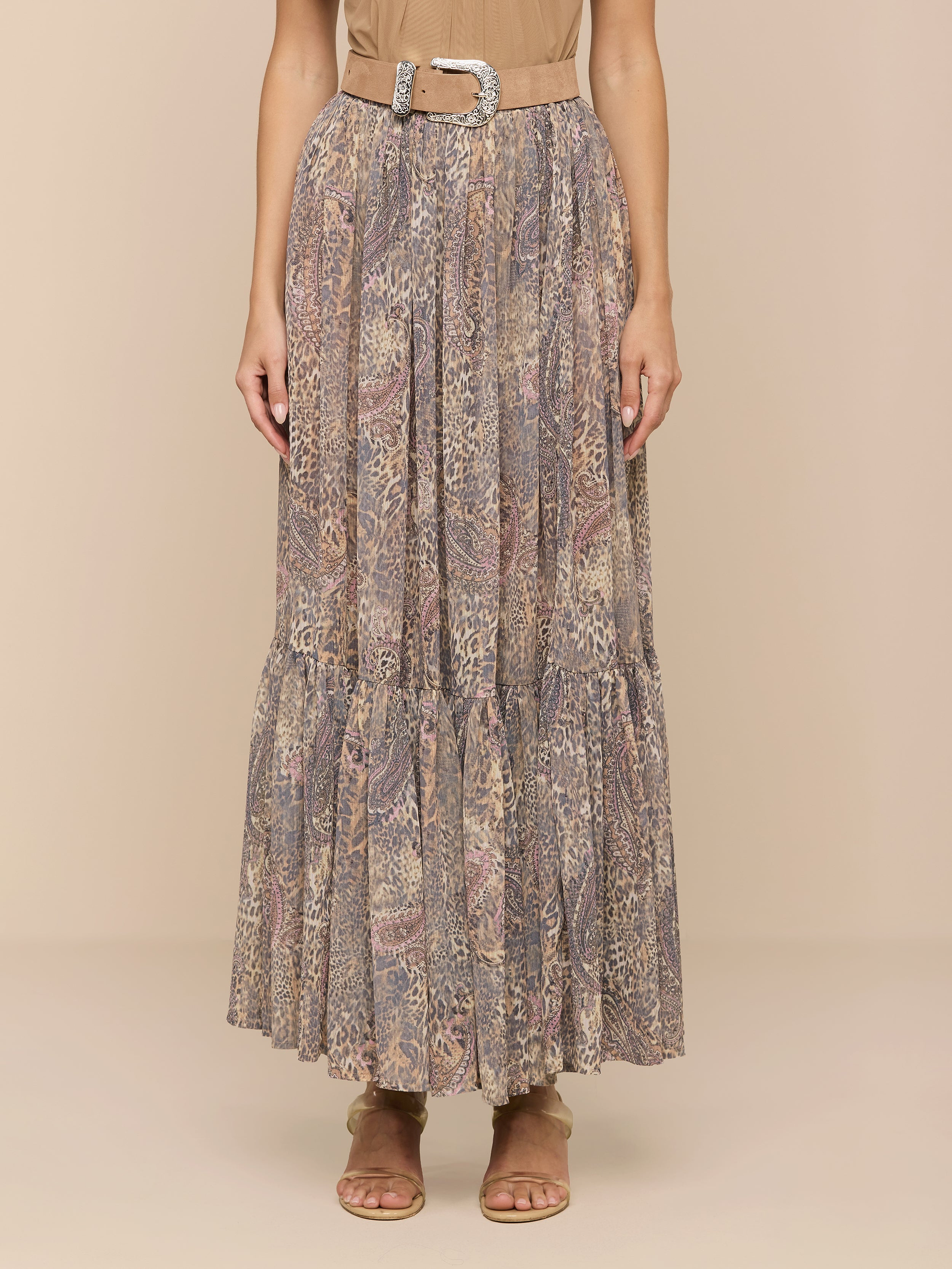 A person wearing a beige top, the Ursie Maxi Skirt—a bohemian paisley leopard print maxi skirt with a belt—and tan sandals stands against a plain light background.