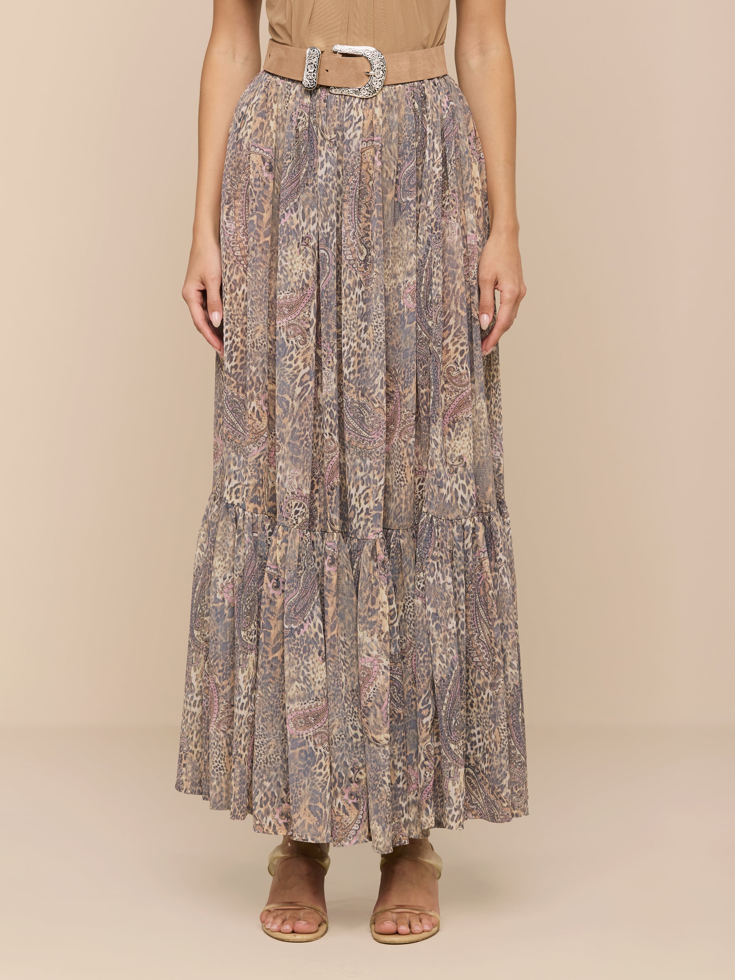 A person wearing a beige top, the Ursie Maxi Skirt—a bohemian paisley leopard print maxi skirt with a belt—and tan sandals stands against a plain light background.