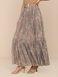 A woman models the Ursie Maxi Skirt—a flowy, ankle-length paisley leopard print design with a belt—paired with a beige top and tan heeled sandals against a plain white background. Her face is not visible in the image.