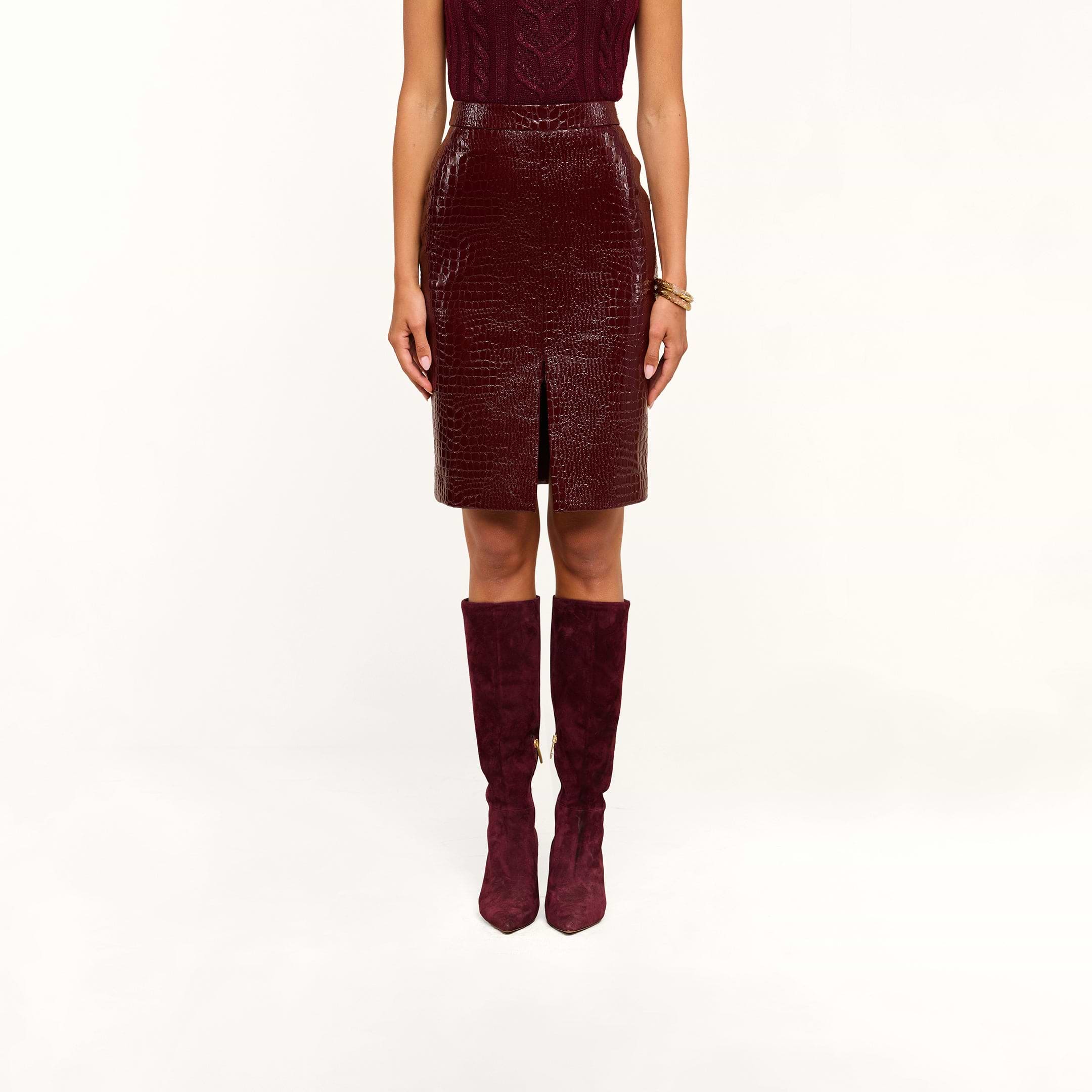 A person wearing a sleeveless burgundy top, the Violetta Vegan Leather Skirt featuring a croc-embossed texture and front slit, and matching burgundy knee-high boots stands against a white background.