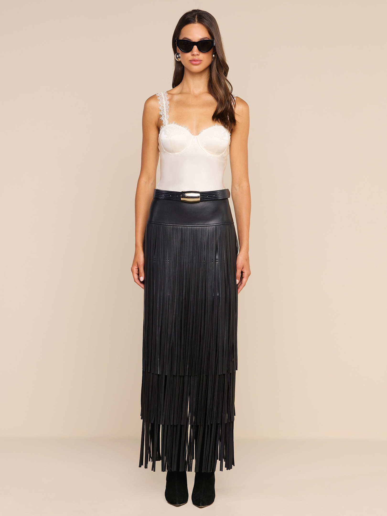 A woman stands against a plain beige background, facing forward with a neutral expression. She wears sunglasses, a white satin camisole with lace straps, and the high-waisted Karoline Fringe Skirt in black vegan leather, featuring long fringe details.