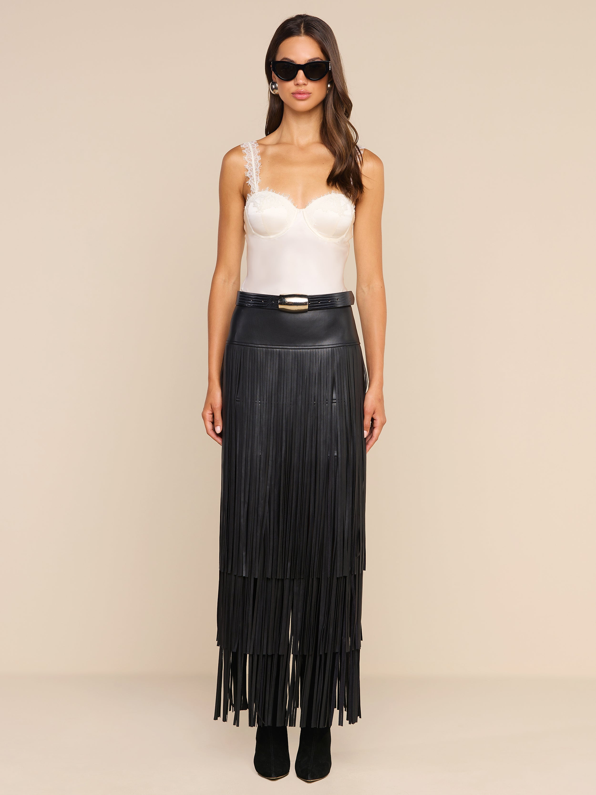 A woman stands against a plain beige background, facing forward with a neutral expression. She wears sunglasses, a white satin camisole with lace straps, and the high-waisted Karoline Fringe Skirt in black vegan leather, featuring long fringe details.