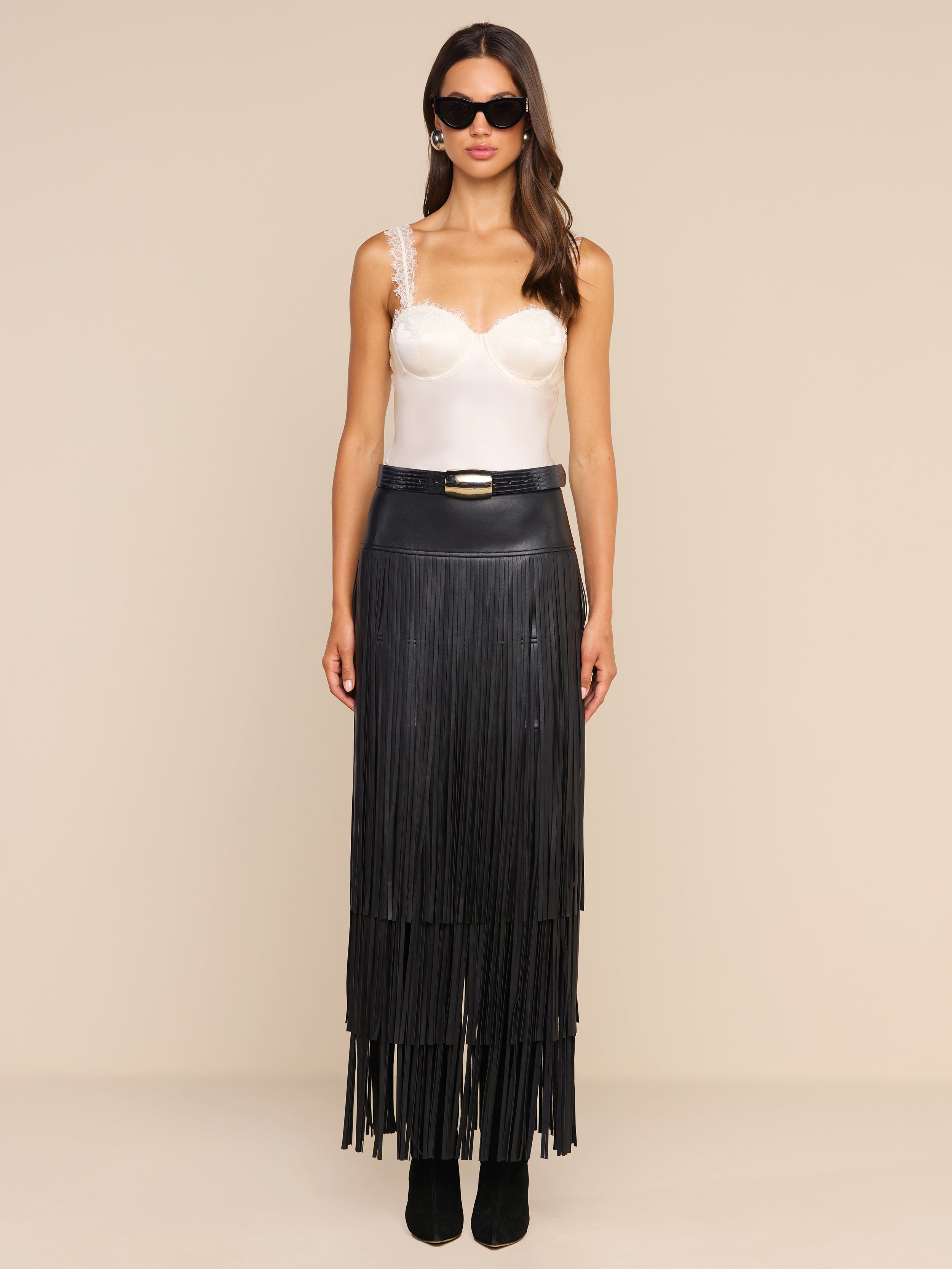 A woman stands against a plain beige background, facing forward with a neutral expression. She wears sunglasses, a white satin camisole with lace straps, and the high-waisted Karoline Fringe Skirt in black vegan leather, featuring long fringe details.