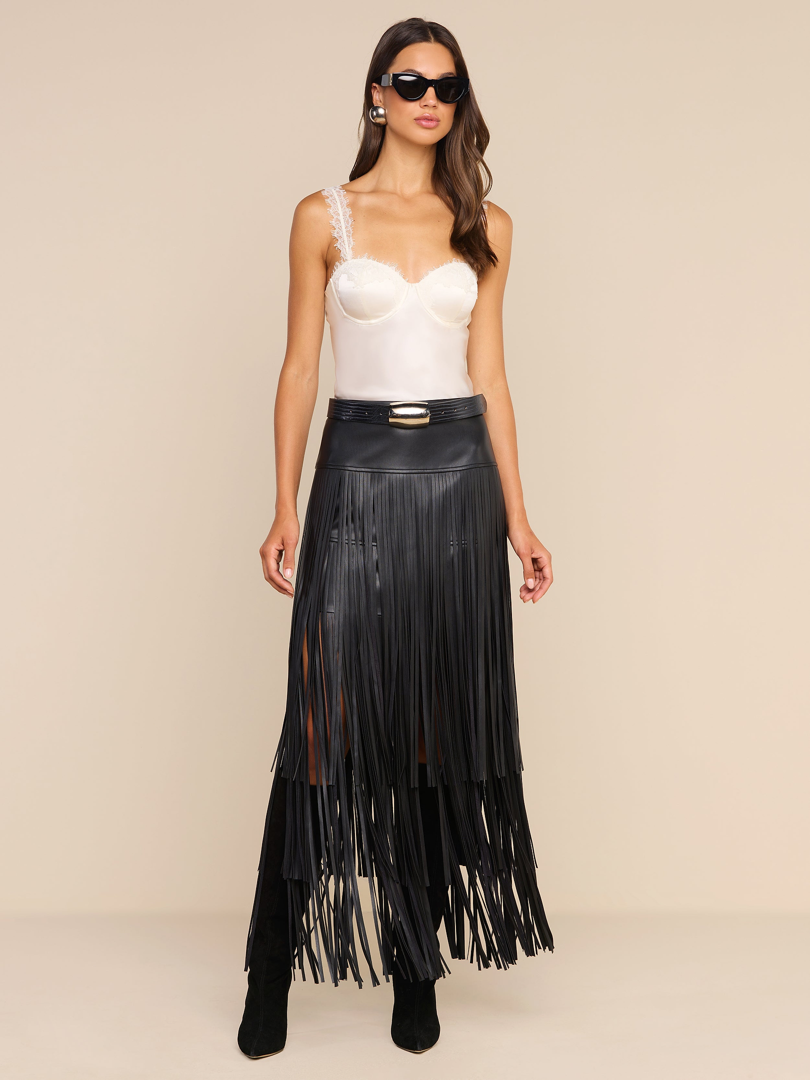 A woman wearing black sunglasses, a white satin camisole, and the Karoline Fringe Skirt with black boots stands against a beige background.