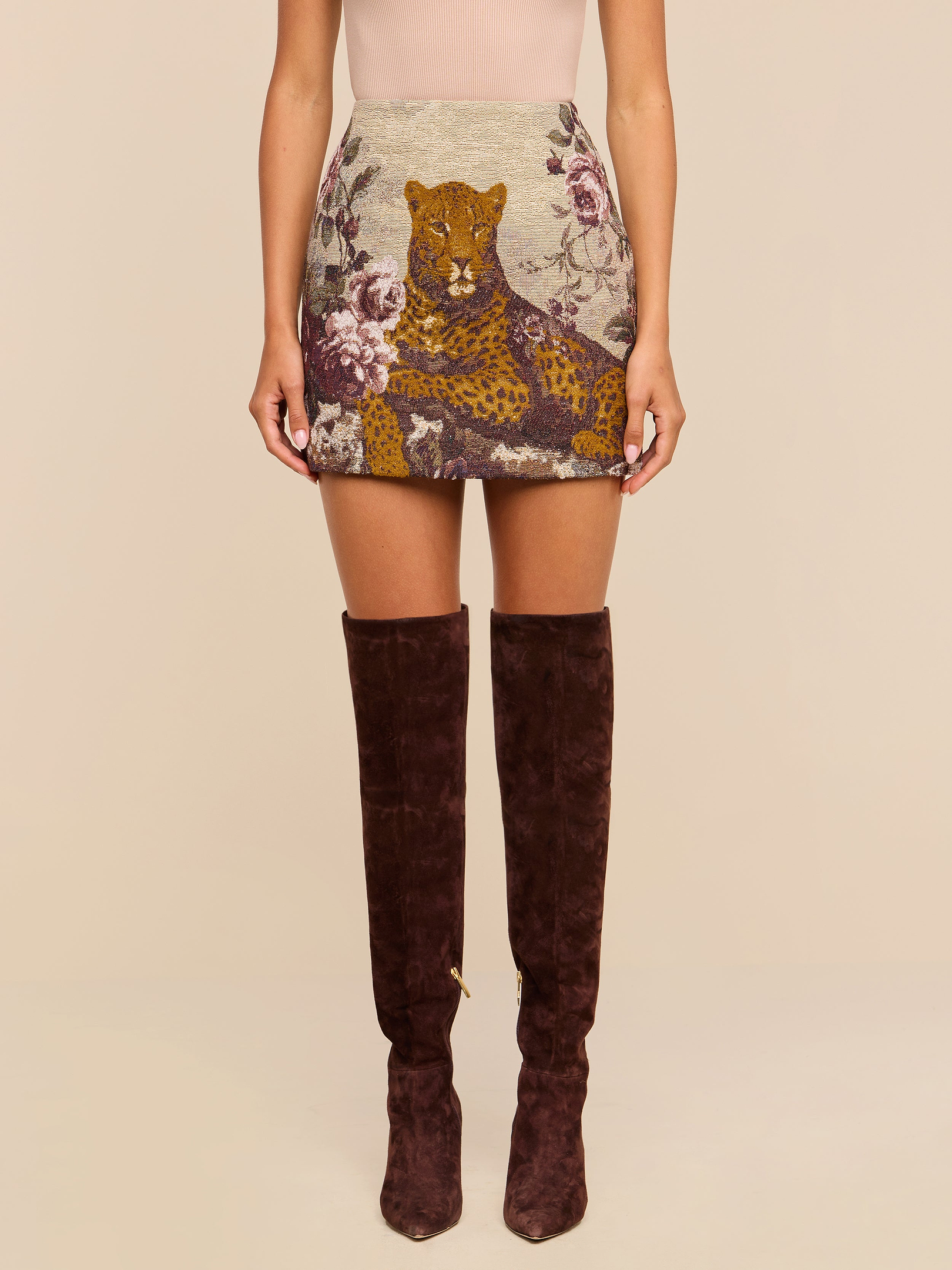 A woman wearing a beige top, the Marseille Mini Skirt—a vintage-inspired mini skirt with a leopard and floral design—and over-the-knee brown suede boots stands against a plain background.
