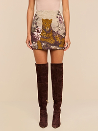 A woman wearing a beige top, the Marseille Mini Skirt—a vintage-inspired mini skirt with a leopard and floral design—and over-the-knee brown suede boots stands against a plain background.