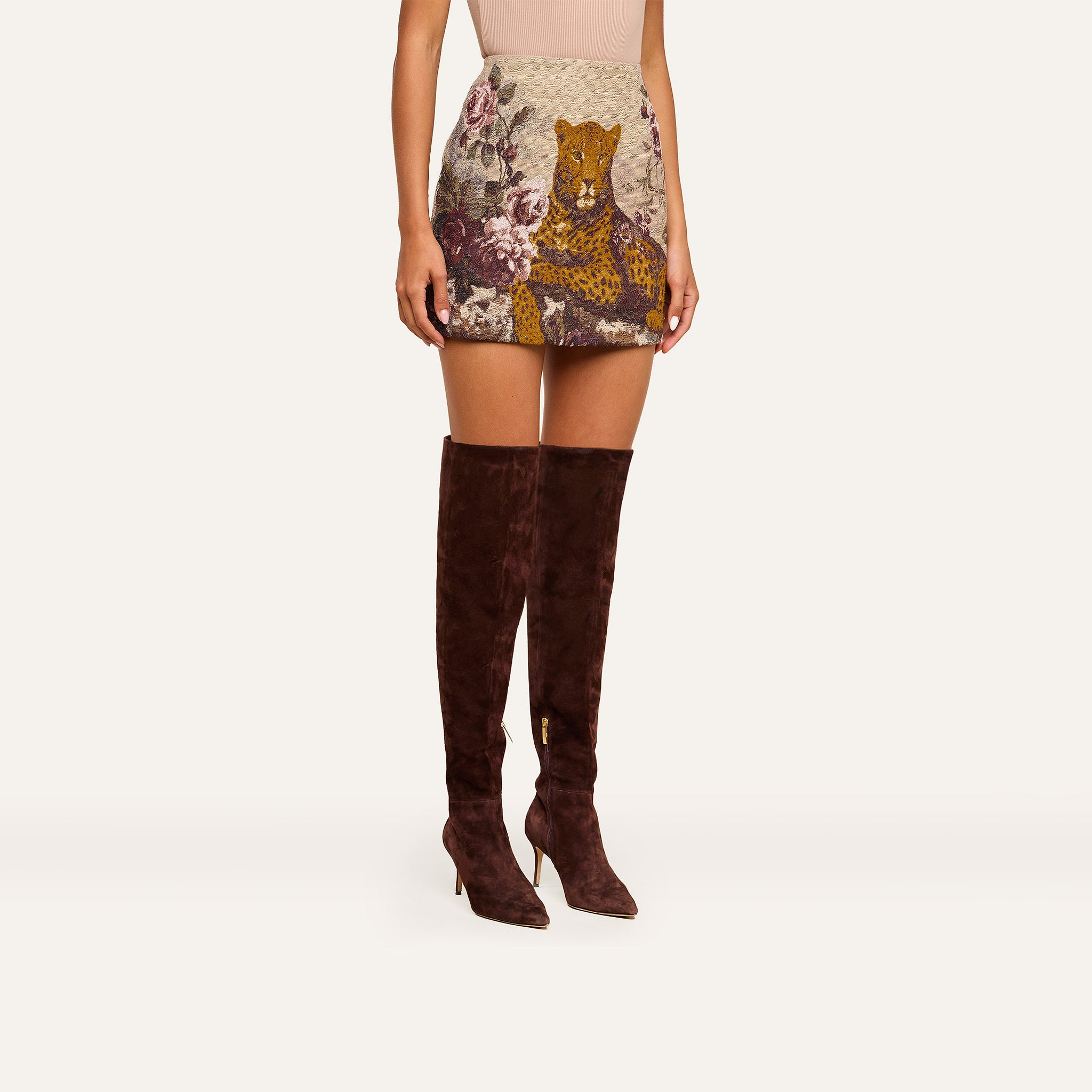 Dressed in the Marseille Mini Skirt, which features a vintage-inspired leopard and floral print, a woman styles her look with dark brown knee-high heeled boots.