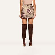 A woman stands facing away, wearing a beige top, the Marseille Mini Skirt with a vintage-inspired purple floral print, and brown suede knee-high boots.