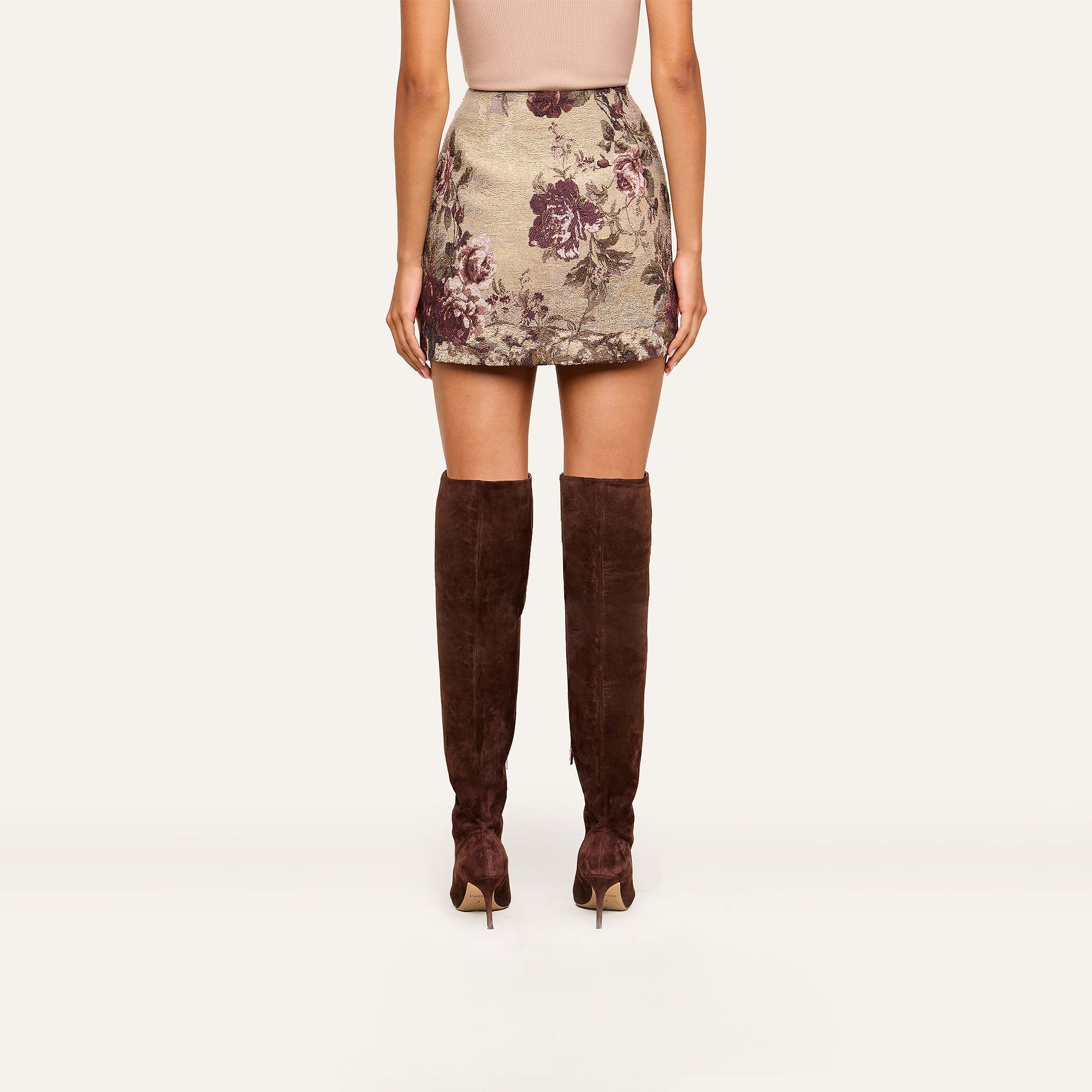 A woman stands facing away, wearing a beige top, the Marseille Mini Skirt with a vintage-inspired purple floral print, and brown suede knee-high boots.