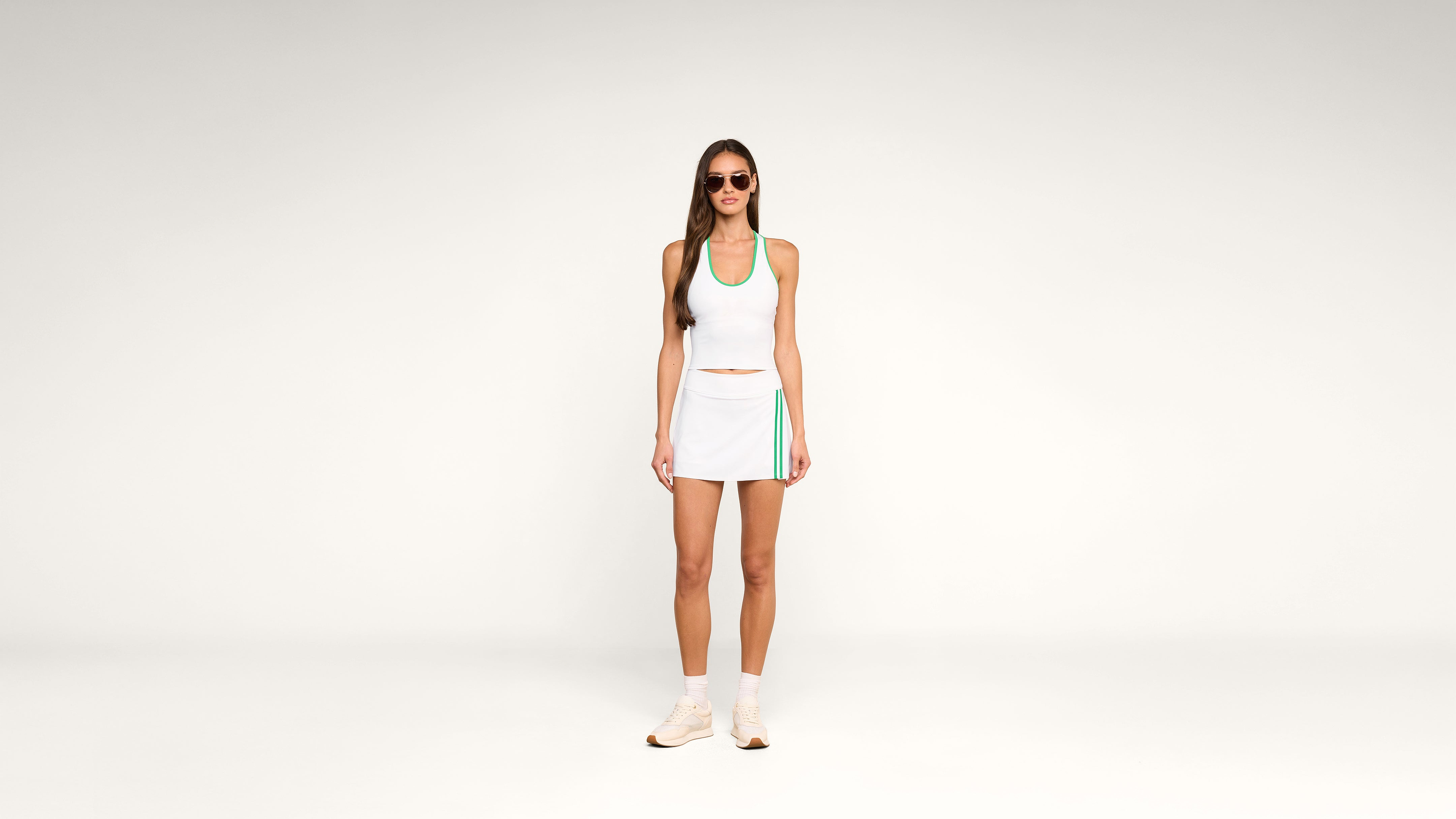 A woman in a white tennis outfit with teal accents, sunglasses, and sneakers stands against a plain white background.