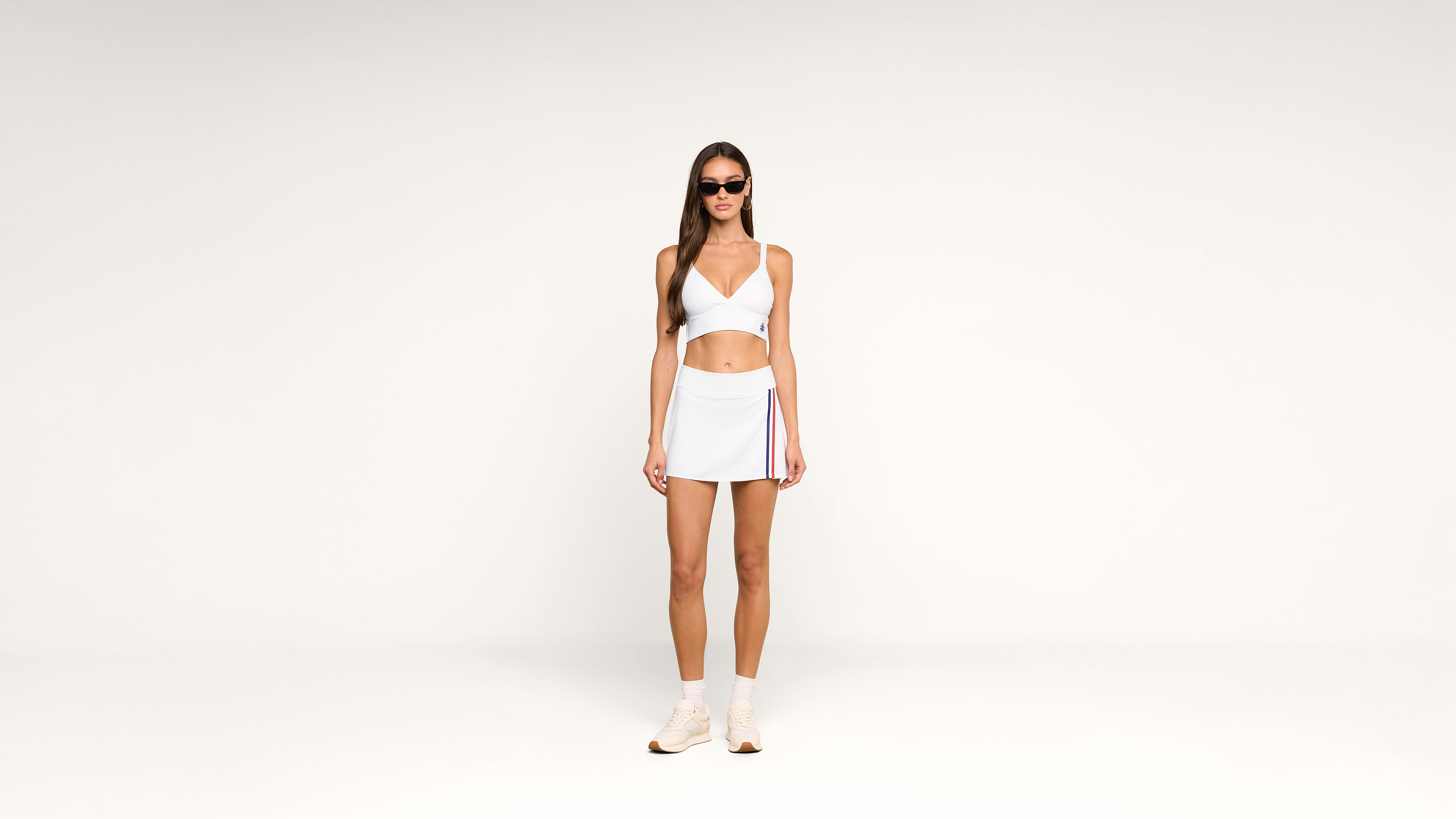 Woman in sunglasses wearing a white crop top, white mini skirt with a side stripe, white socks, and sneakers stands against a plain white background.