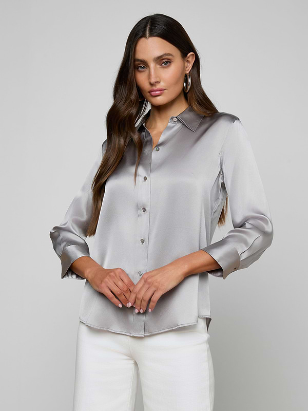 Woman with long brown hair wearing the Dani Silk Blouse and white pants, standing against a plain light background, looking at the camera with hands gently clasped.