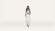 A woman wearing a shiny silver blouse, white wide-leg pants, and beige heels stands against a plain white background, looking to the side.