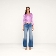 A woman stands facing forward against a plain white background, wearing the shiny lavender Dani Silk Blouse in silk charmeuse, blue high-waisted wide-leg jeans, a brown belt, and beige heels.
