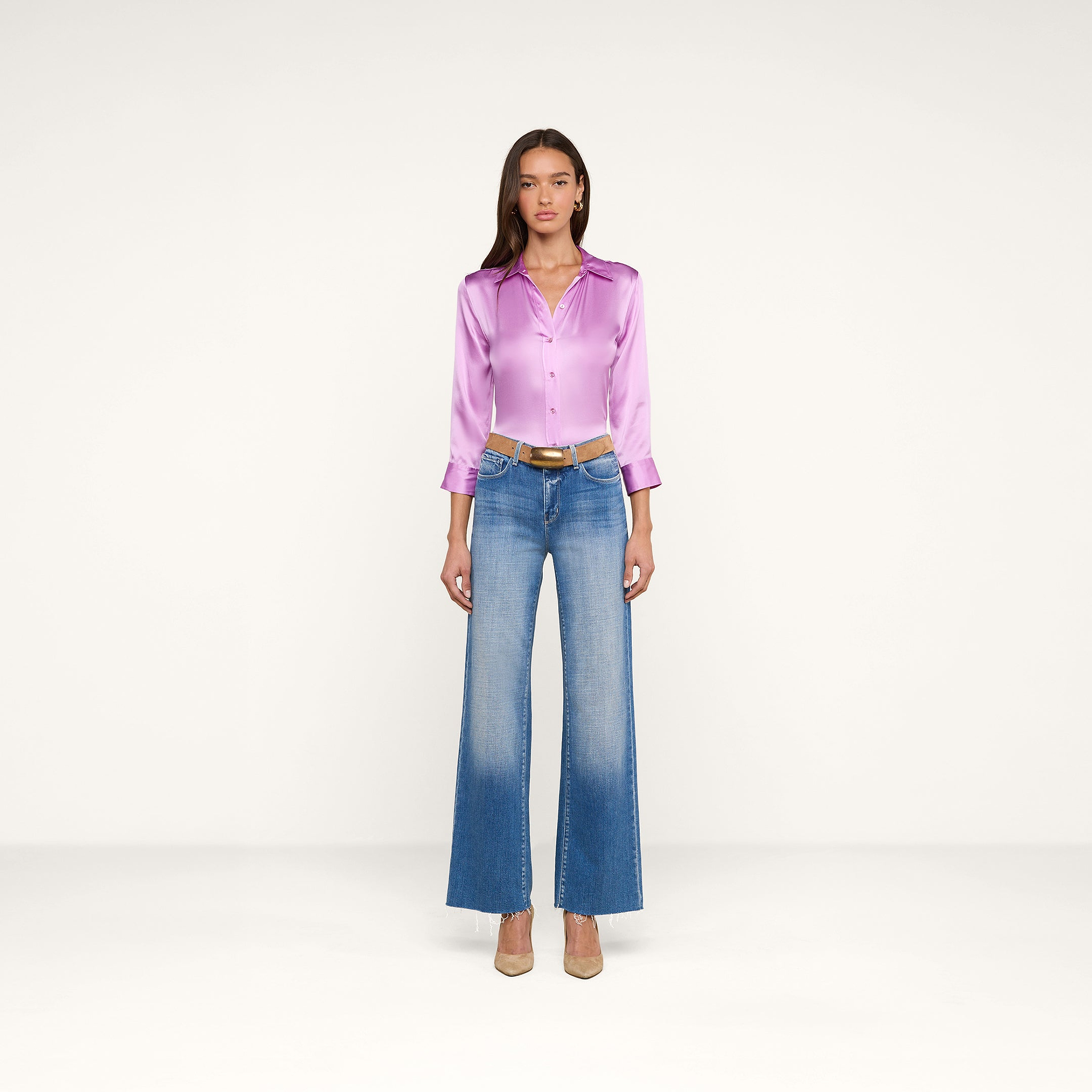 A woman stands facing forward against a plain white background, wearing the shiny lavender Dani Silk Blouse in silk charmeuse, blue high-waisted wide-leg jeans, a brown belt, and beige heels.