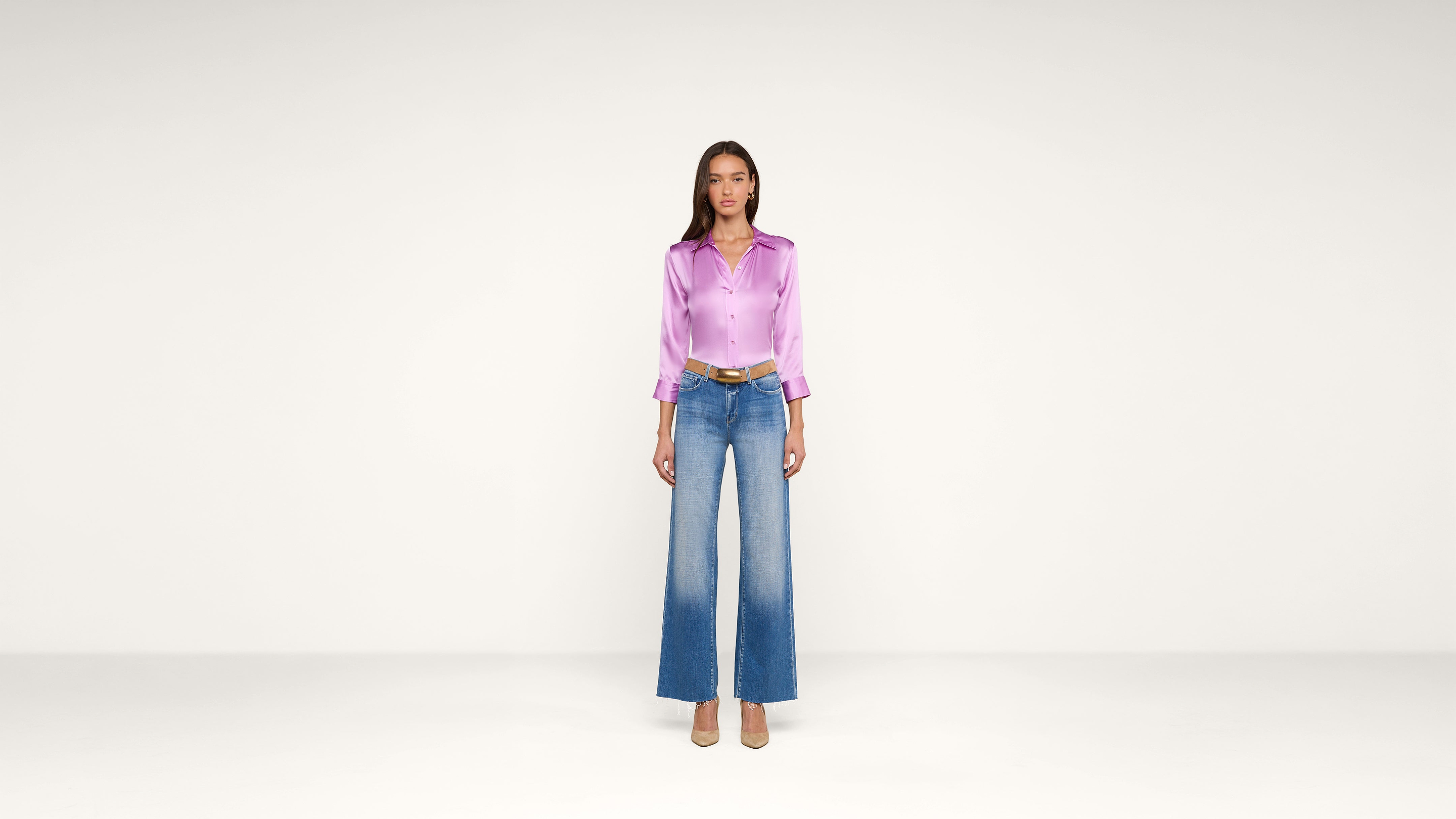 Woman standing against a plain white background wearing a shiny lavender blouse, wide-leg blue jeans with a faded effect, a brown belt, and beige heels.
