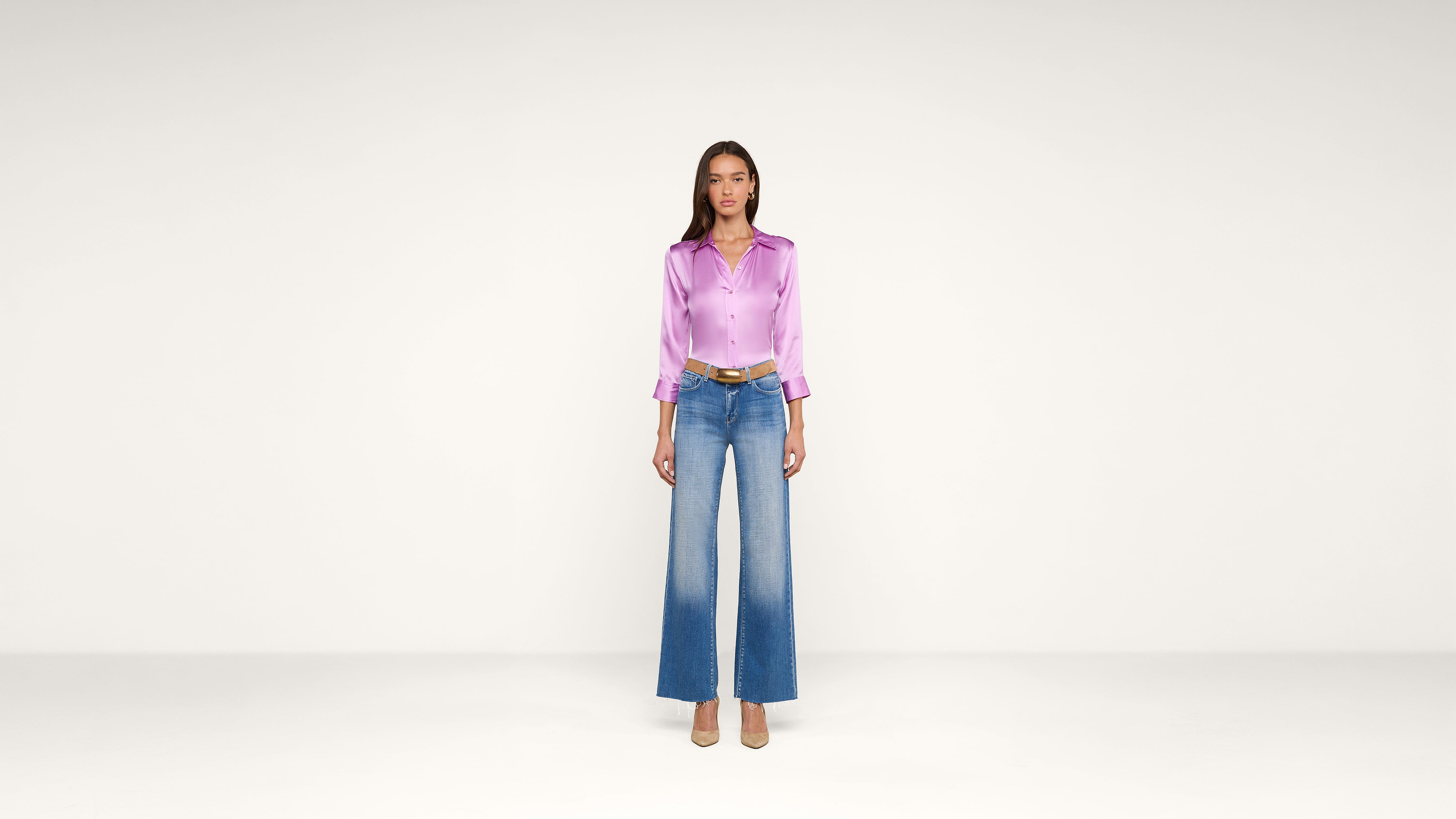 Woman standing against a plain white background wearing a shiny lavender blouse, wide-leg blue jeans with a faded effect, a brown belt, and beige heels.