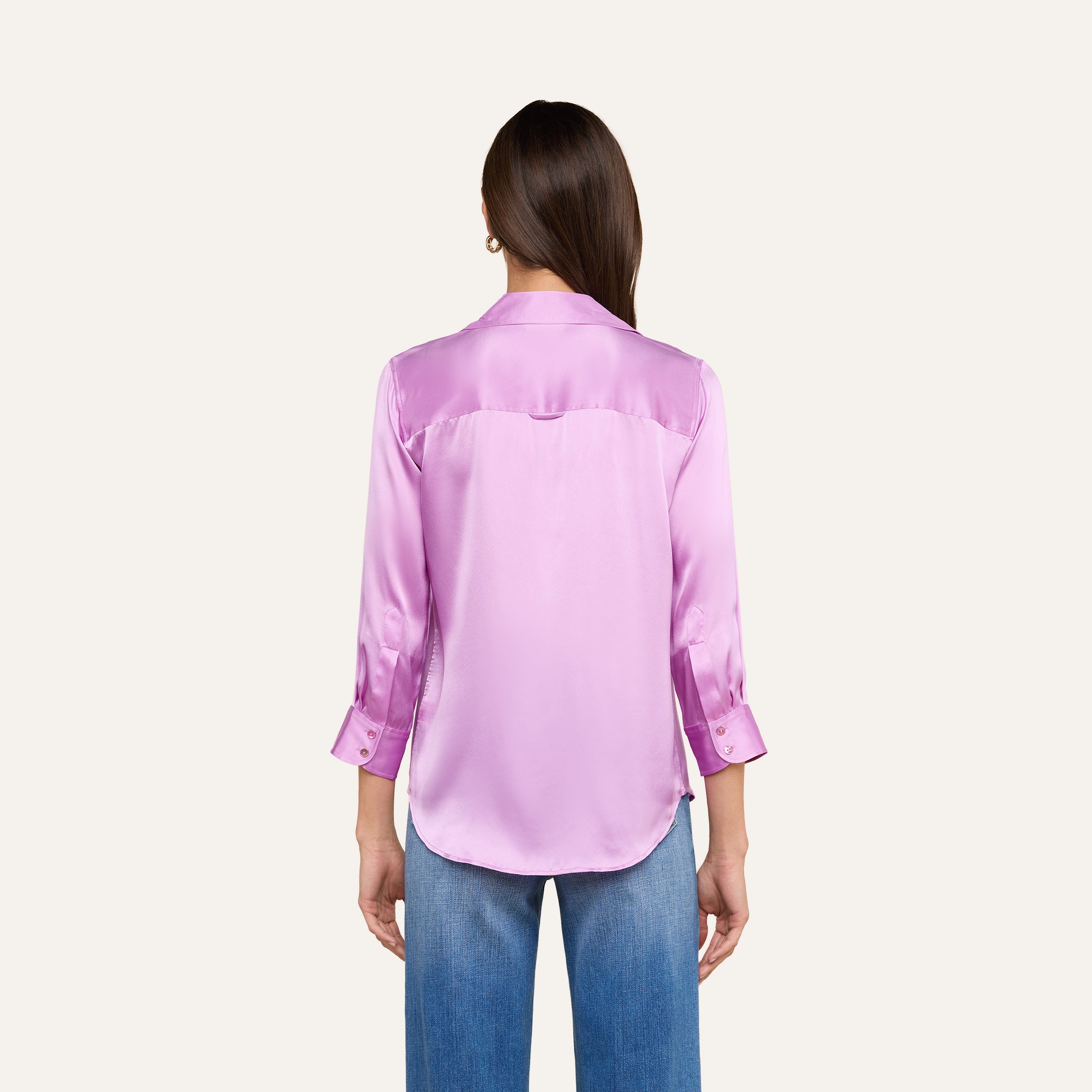 A person with long brown hair, wearing the Dani Silk Blouse in shiny, light purple silk charmeuse with three-quarter sleeves, stands facing away. They are also wearing blue jeans.