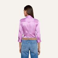 A woman stands with her back to the camera, wearing the Dani Silk Blouse in shiny pink charmeuse with three-quarter sleeves, paired with blue jeans. The background is plain and light-colored.
