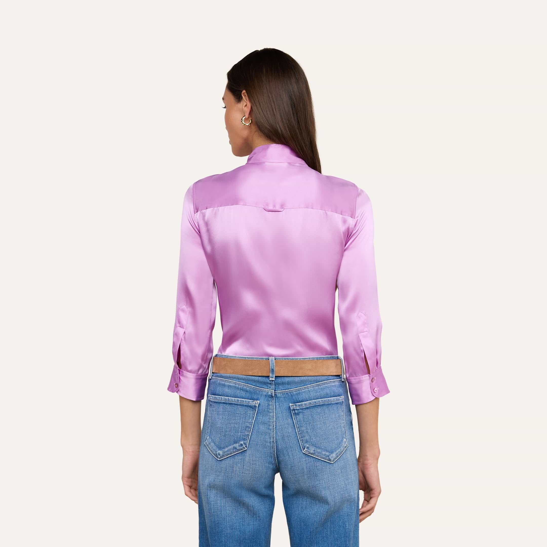 A woman stands with her back to the camera, wearing the Dani Silk Blouse in shiny pink charmeuse with three-quarter sleeves, paired with blue jeans. The background is plain and light-colored.
