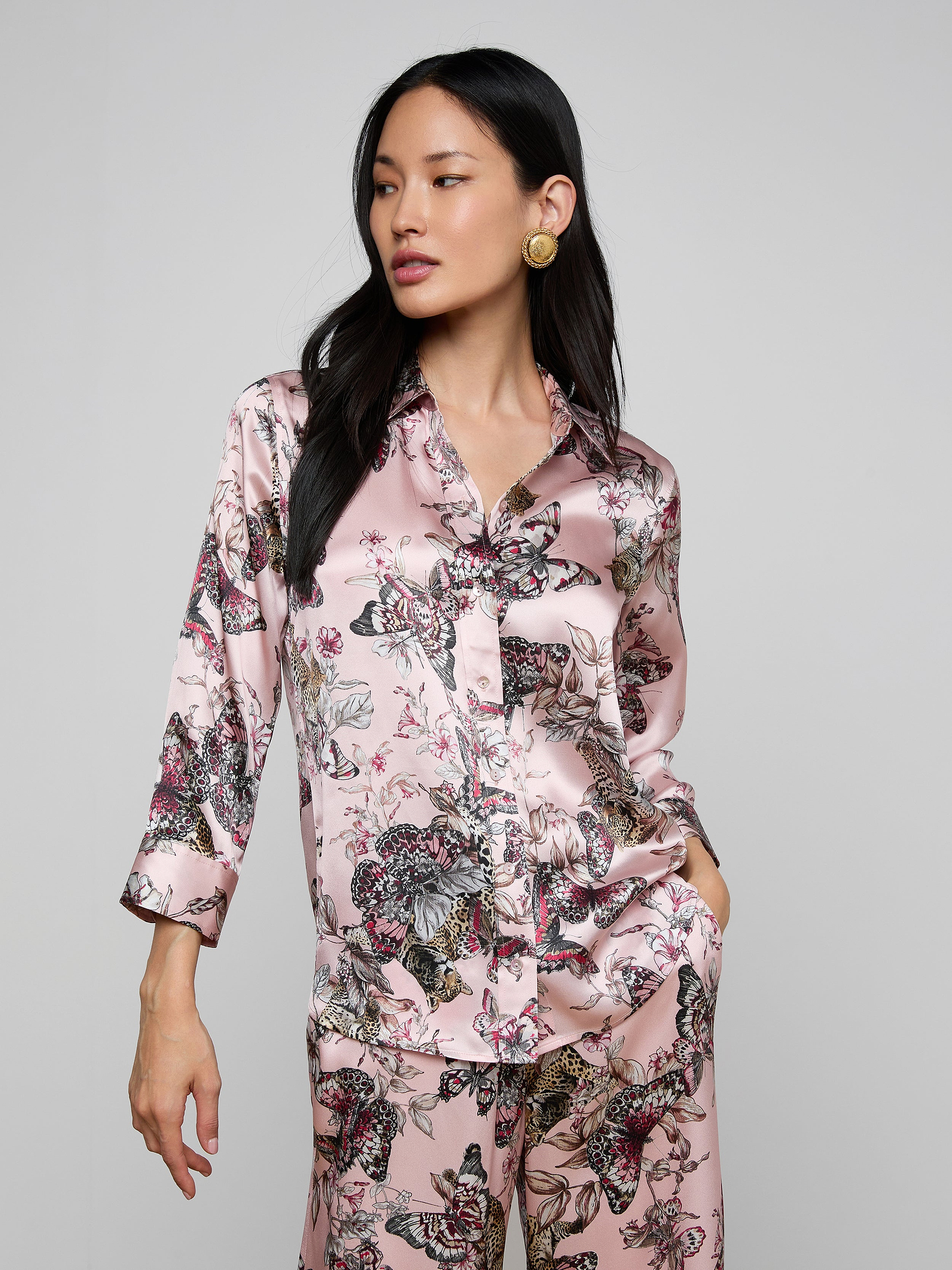 A woman wearing the Dani Silk Blouse in a shiny, light pink silk fabric with a butterfly and floral pattern poses with one hand in her pocket against a plain light background.