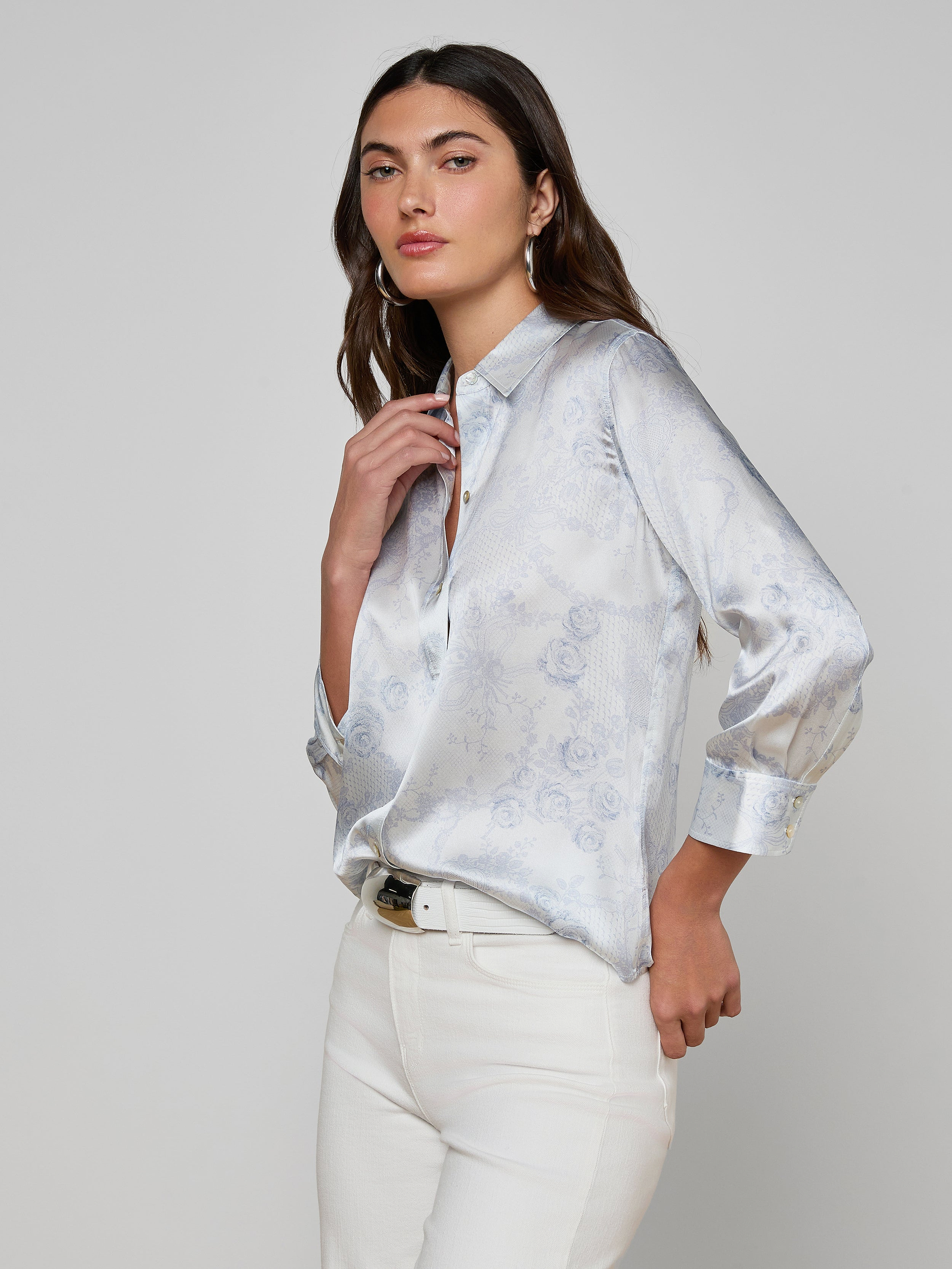 A woman with long brown hair wears the Dani Silk Blouse—a light blue satin button-down featuring subtle floral patterns—paired with white pants against a plain, off-white background.