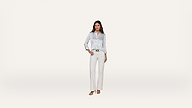 A woman stands against a plain white background, wearing a light blue button-up shirt, white pants, and sandals. She has one hand in her pocket and looks confidently at the camera.