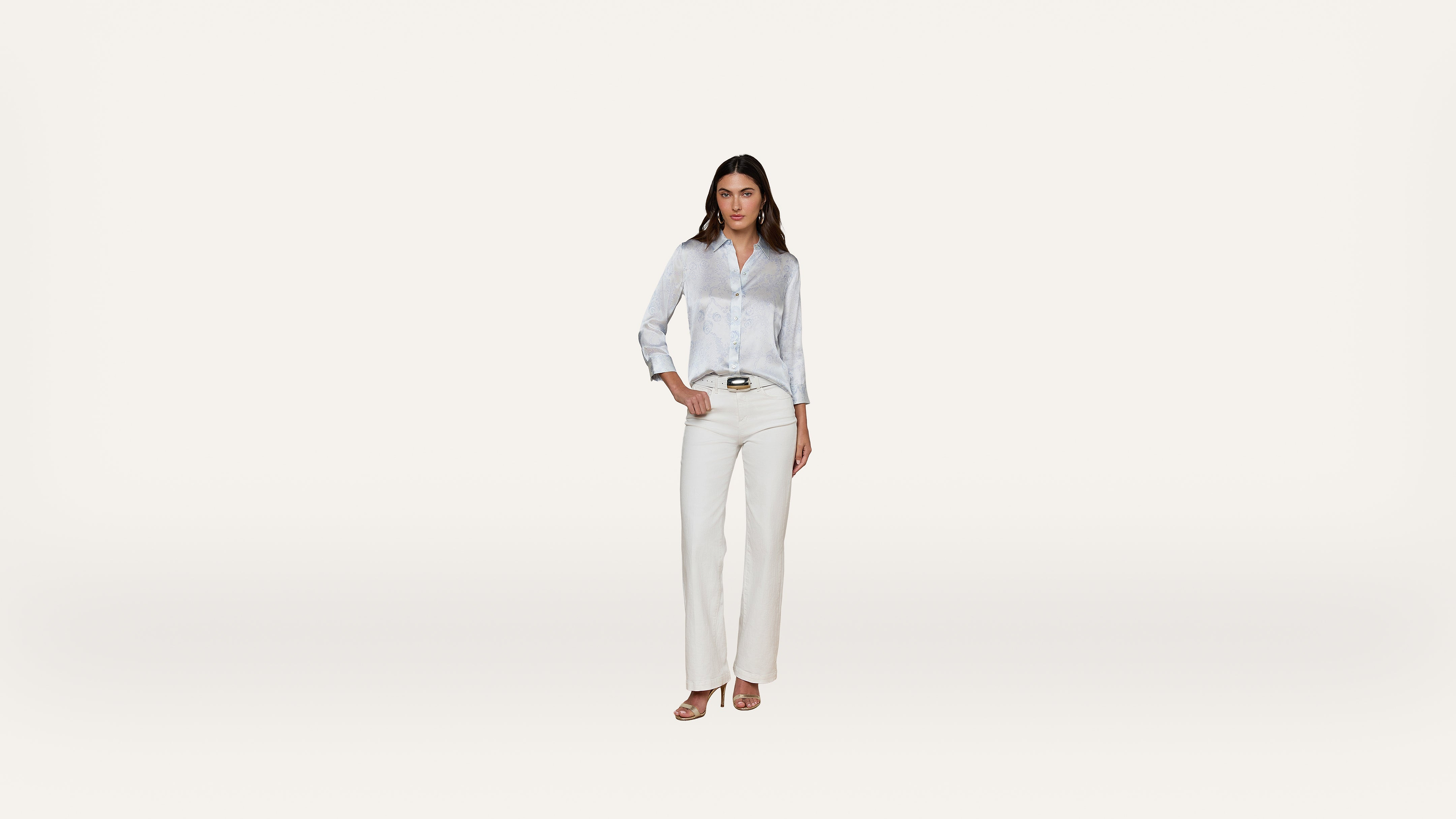 A woman stands against a plain white background, wearing a light blue button-up shirt, white pants, and sandals. She has one hand in her pocket and looks confidently at the camera.