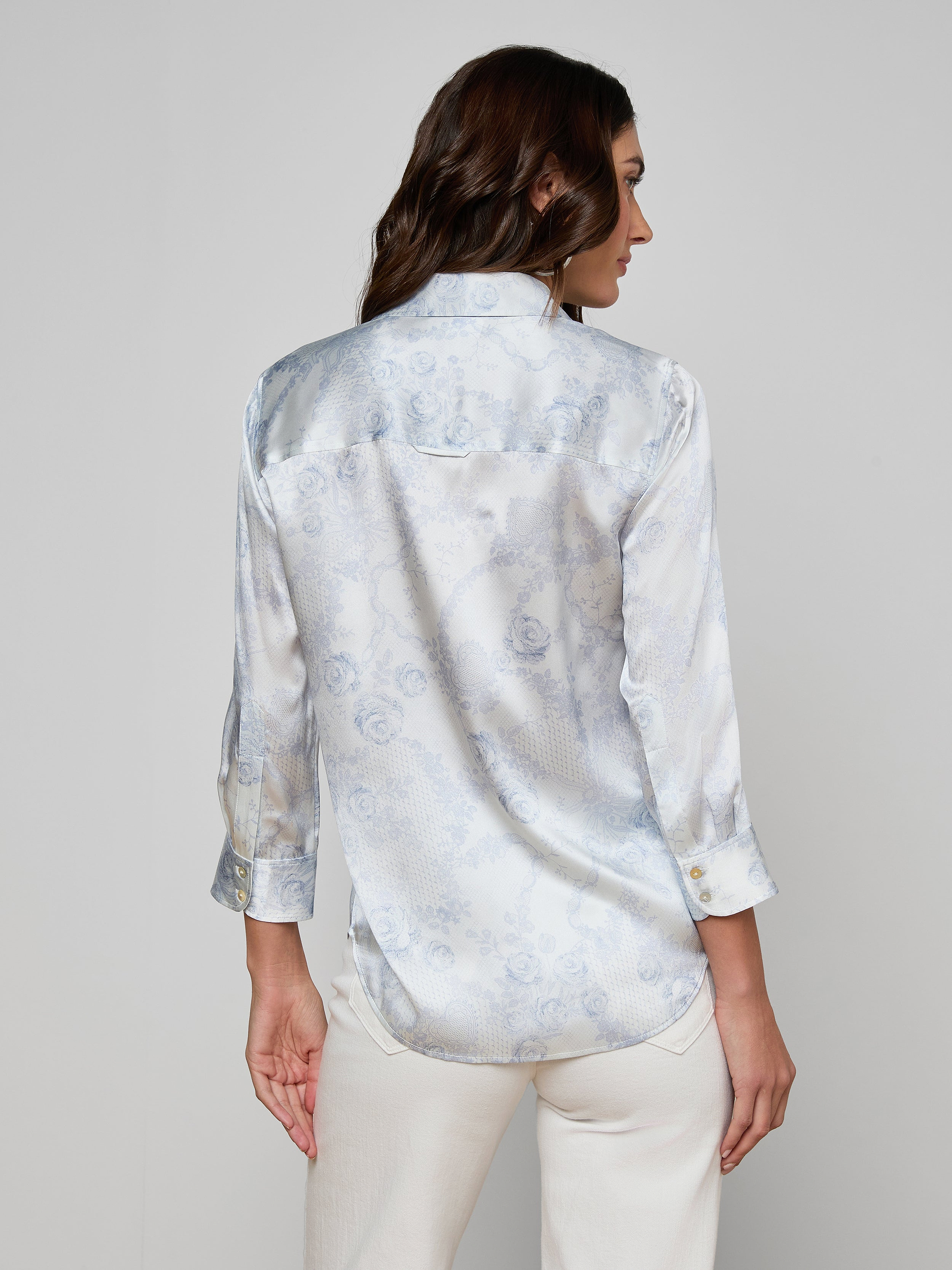 A woman with long brown hair, wearing the Dani Silk Blouse—a light blue patterned silk button-up shirt with rolled-up sleeves—and white pants, stands with her back to the camera against a plain background.