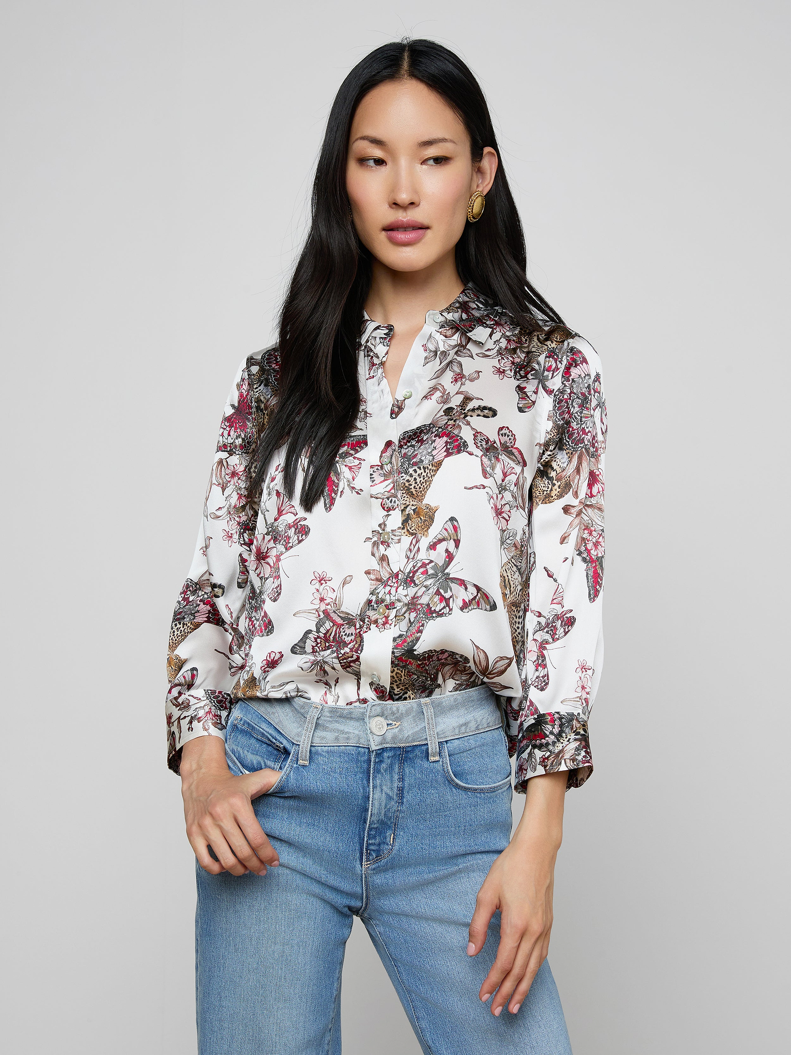 A woman wearing the Dani Silk Blouse—a patterned white top featuring floral, bird, and butterfly jungle print designs—paired with blue jeans stands against a plain background, looking slightly to the side with one hand in her pocket.