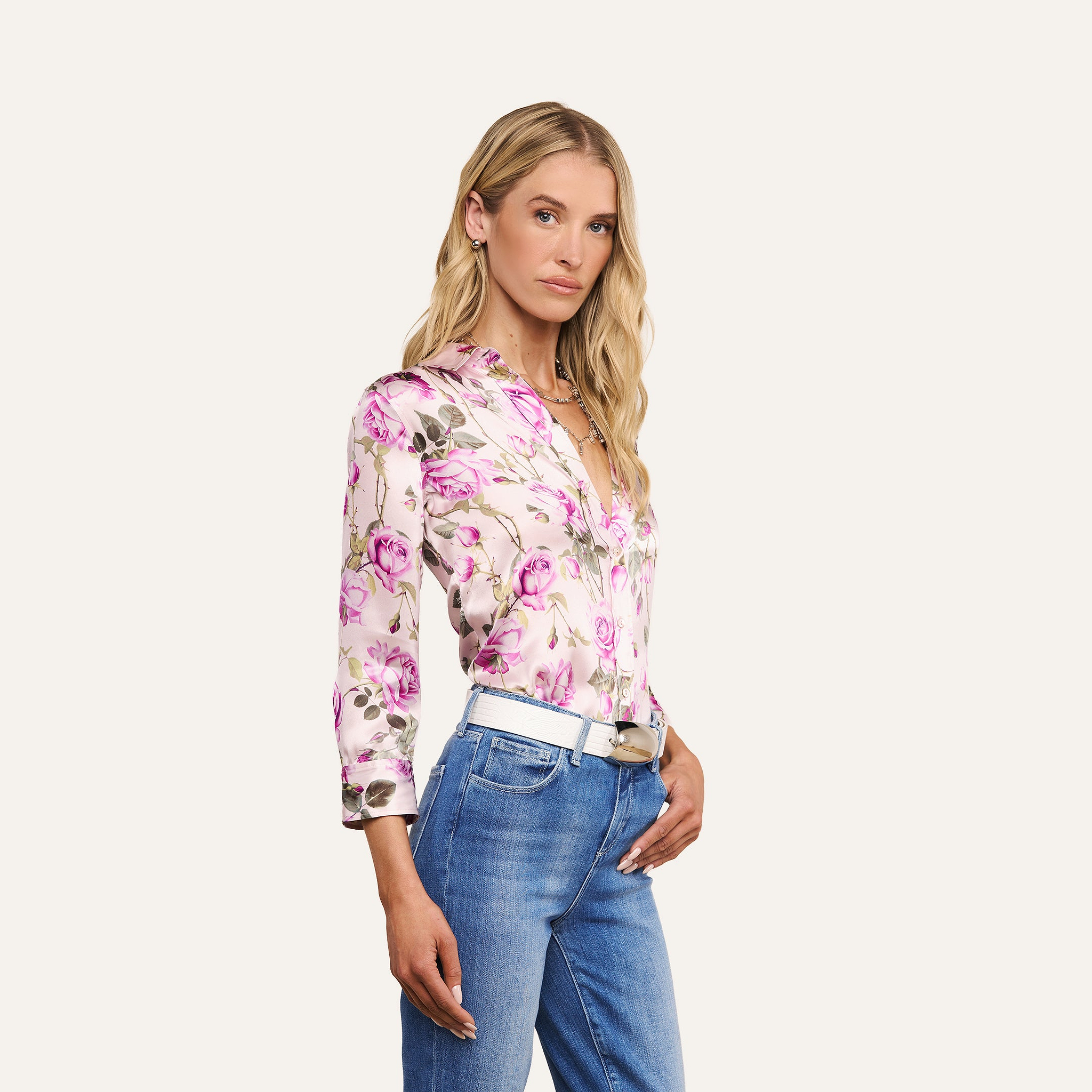 A woman with long blonde hair wears a pink rose print Dani Silk Blouse and blue jeans with a white belt, standing against a plain light background.