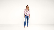 A woman stands against a plain white background, wearing a pink floral blouse, blue flared jeans, and sandals. She has blonde hair and a neutral expression, with her arms relaxed by her sides.