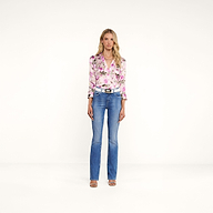 A woman with long blonde hair and a neutral expression stands against a plain white background, wearing the Dani Silk Blouse featuring a rose print, paired with blue jeans, a white belt, and open-toe heels.