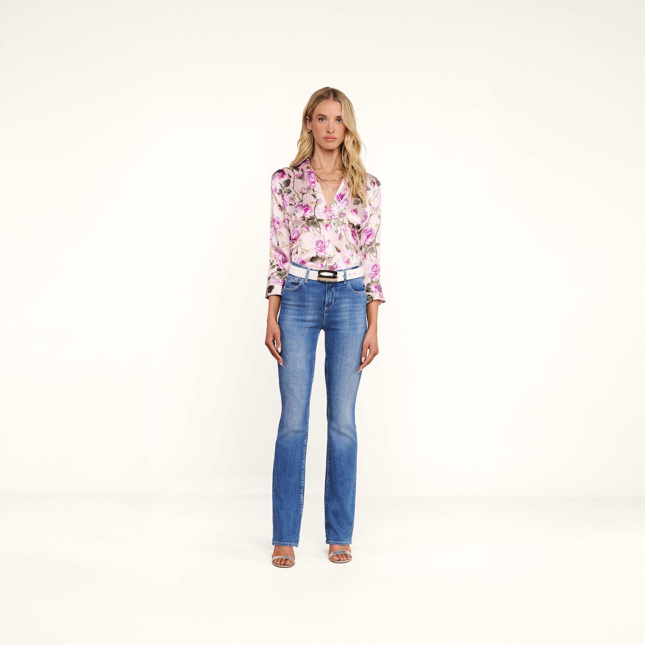 A woman with long blonde hair and a neutral expression stands against a plain white background, wearing the Dani Silk Blouse featuring a rose print, paired with blue jeans, a white belt, and open-toe heels.