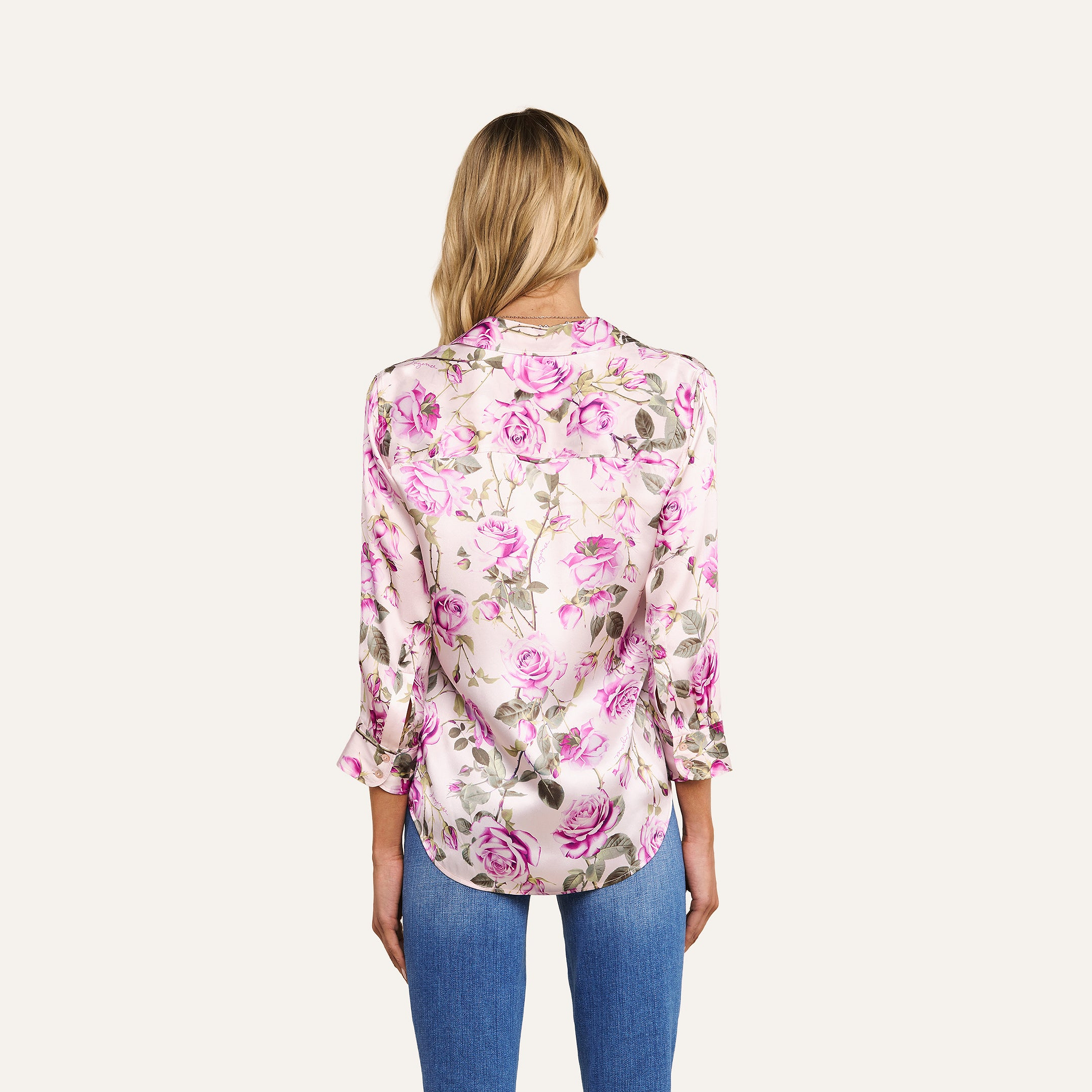 A woman with long blonde hair stands with her back to the camera against a plain light background, wearing the Dani Silk Blouse in a pink rose print paired with blue jeans.