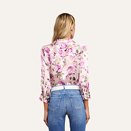 A woman with long blonde hair is seen from behind, wearing a rose print Dani Silk Blouse with rolled-up sleeves and high-waisted blue jeans against a plain, light background.