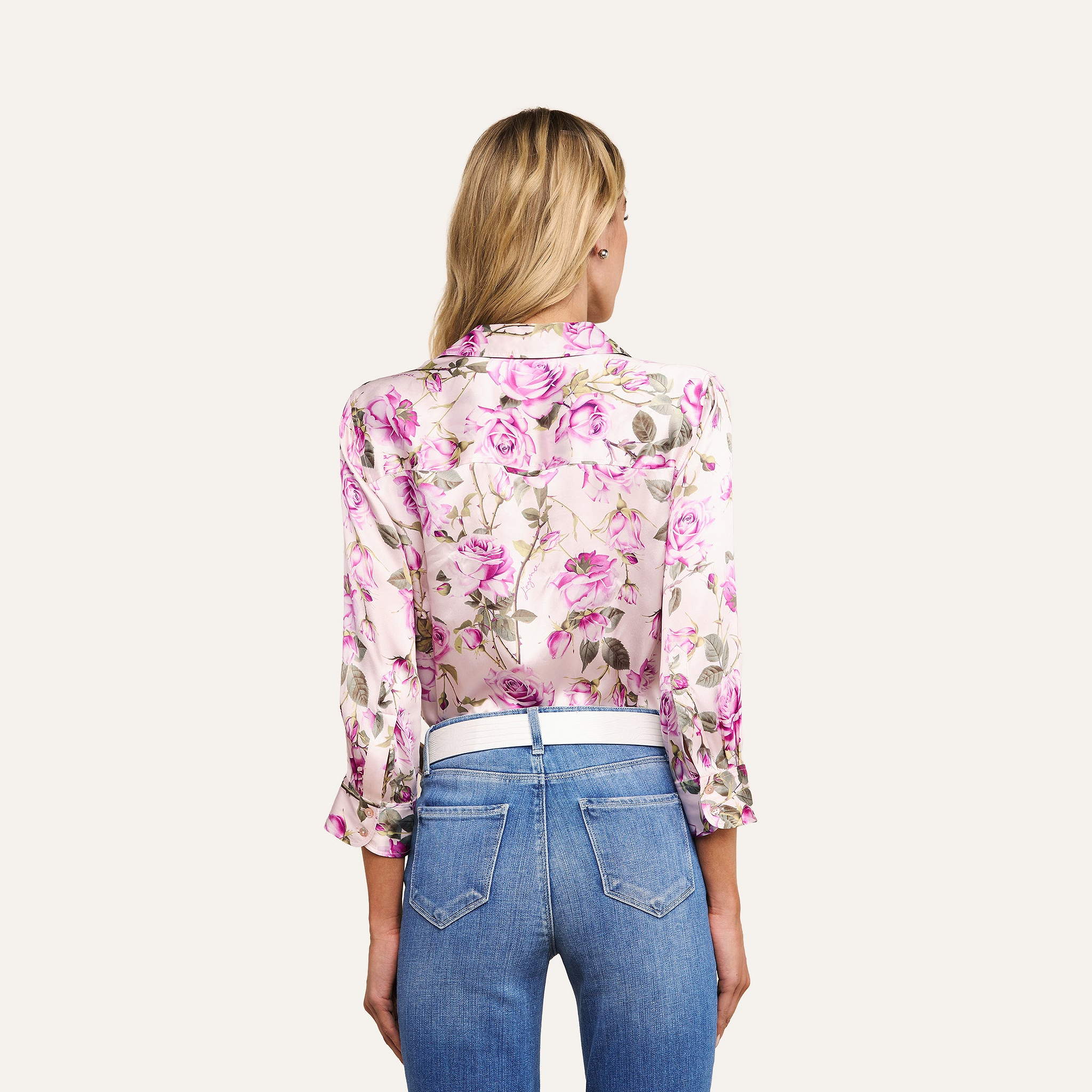 A woman with long blonde hair is seen from behind, wearing a rose print Dani Silk Blouse with rolled-up sleeves and high-waisted blue jeans against a plain, light background.