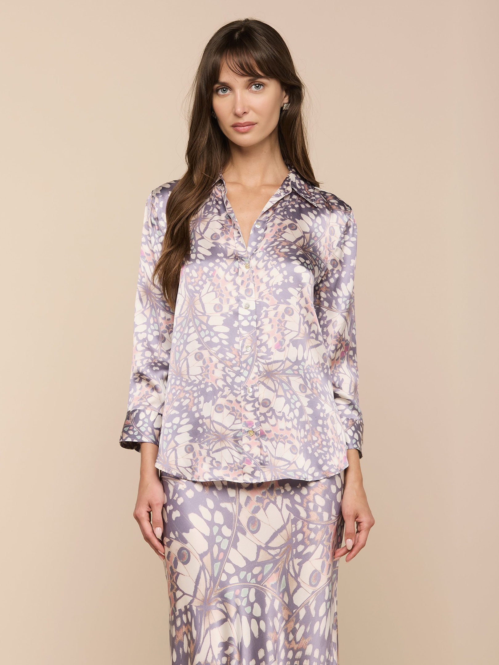 A woman wears the Dani Silk Blouse—a long-sleeve, button-down shirt in silk—paired with a matching skirt. Both pieces showcase a light purple and beige abstract floral pattern reminiscent of butterfly wings, as she stands against an off-white background.